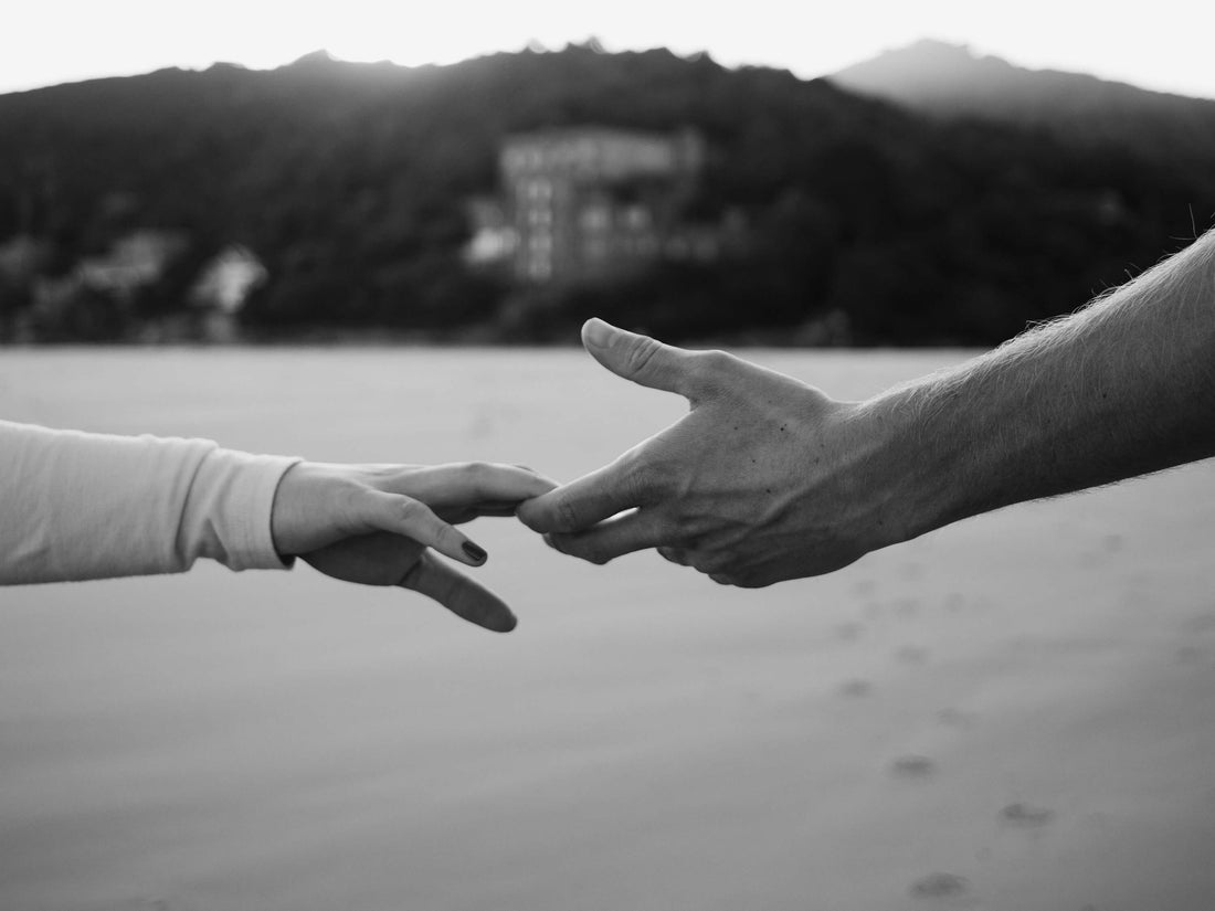 Two hands reach toward each other over a calm body of water with distant hills, captured in black and white