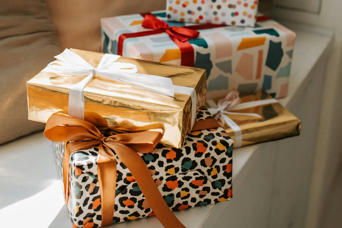 Assorted wrapped gifts stacked on a surface, including a shiny gold box with a white ribbon