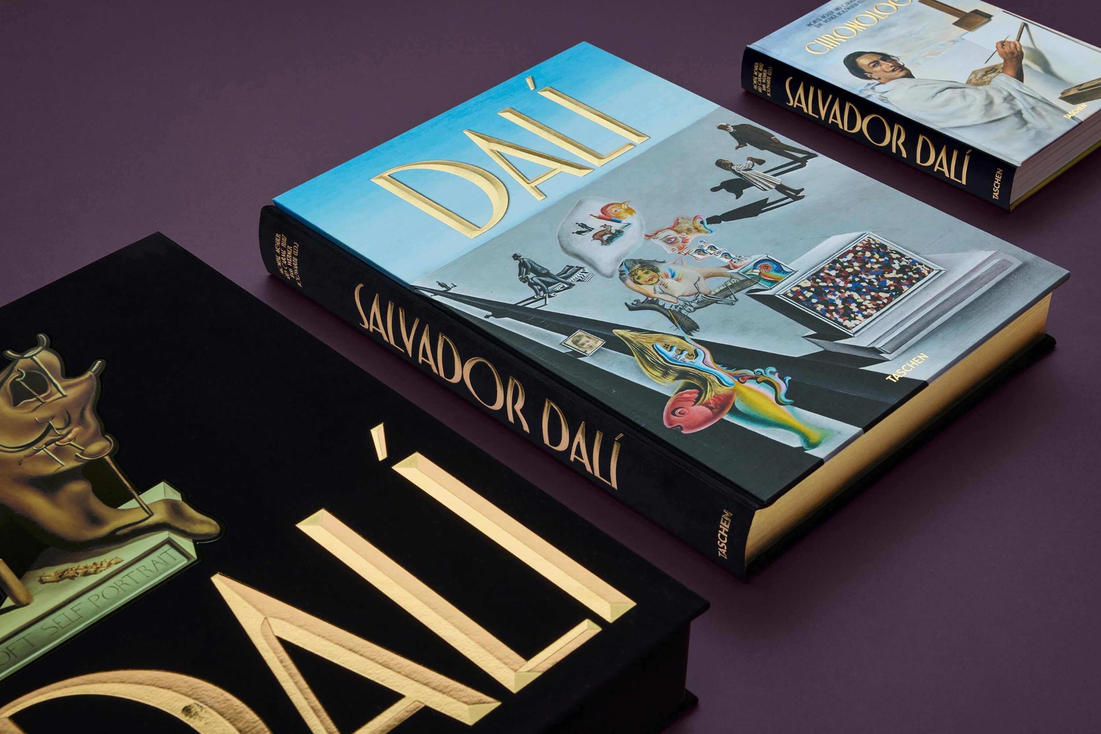 Dalí art books stacked on a purple surface, one with a bright blue cover and gold title alongside another smaller Salvador Dalí volume.