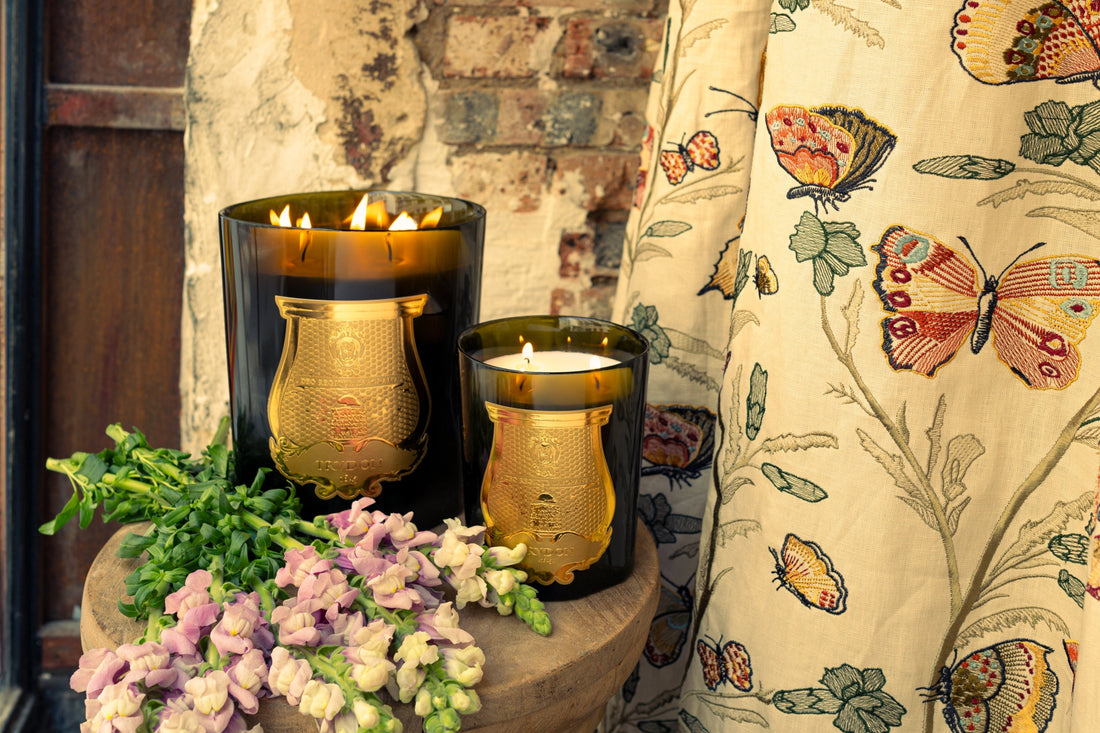 Two dark glass candle holders with gold plaques glow beside greenery and pale purple flowers against a rustic brick backdrop.