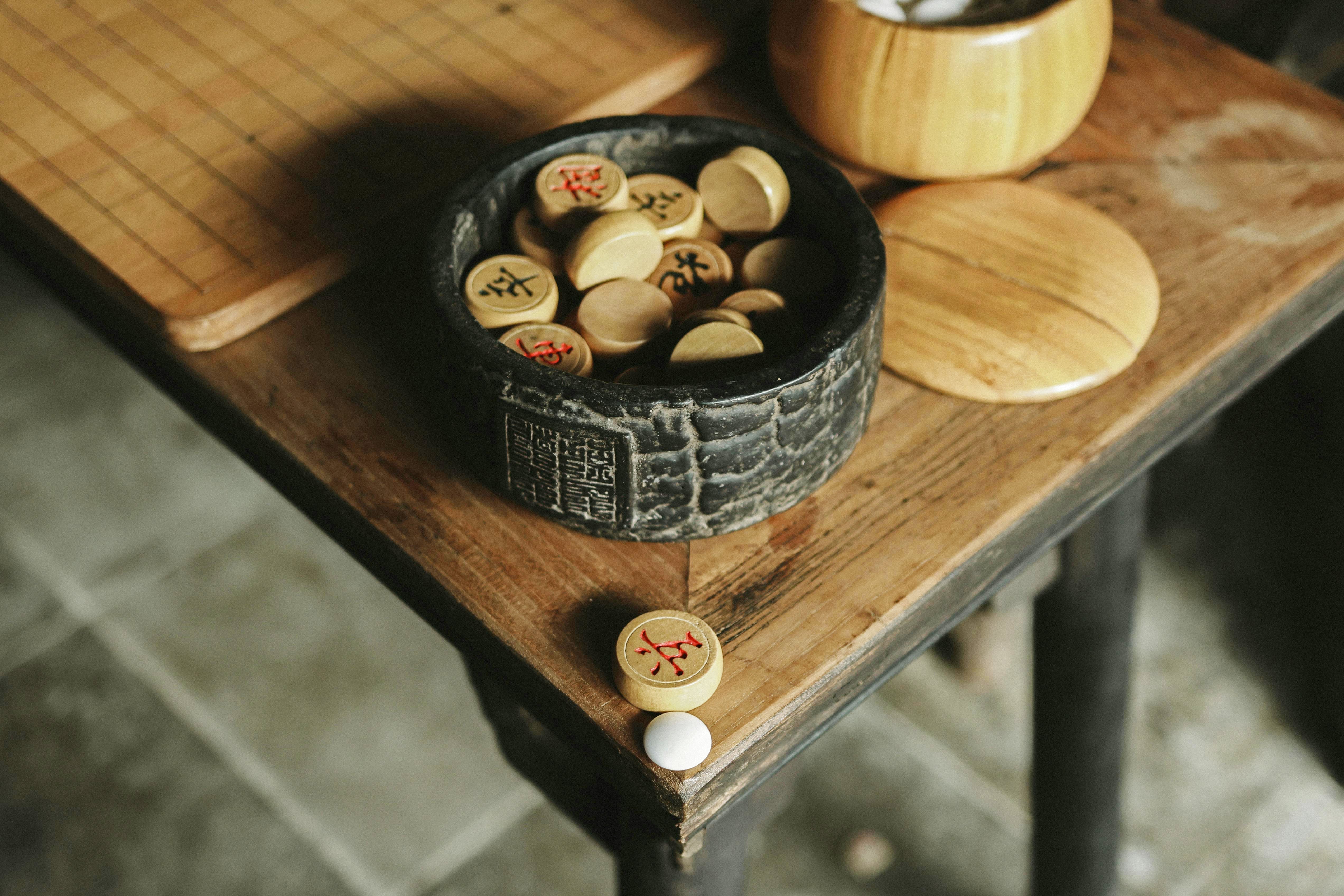 Xiangqi chess pieces inside a round black container on a wooden table with a few pieces spilled nearby