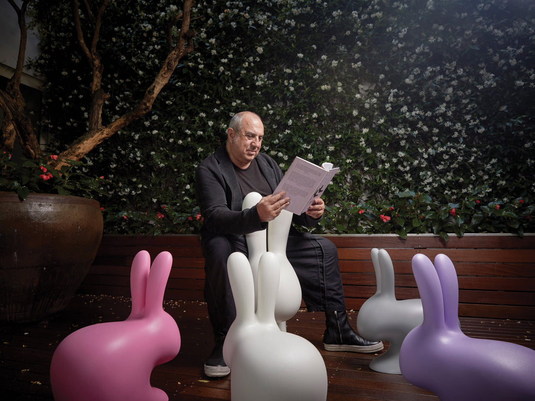 Man sits outdoors reading a book among pastel rabbit sculpture figures on a wooden deck with greenery in the background.