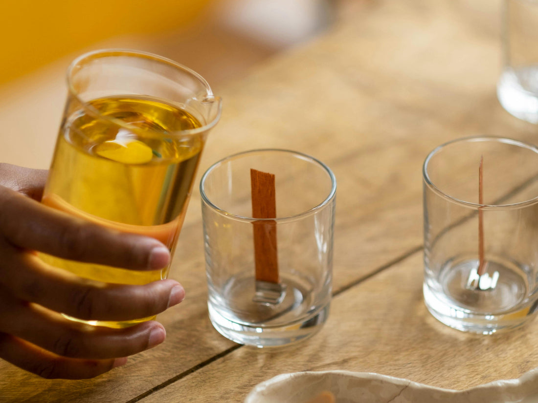 Hand holds a glass of golden beverage with a lemon slice, beside two clear glasses with cinnamon sticks on a wooden table.