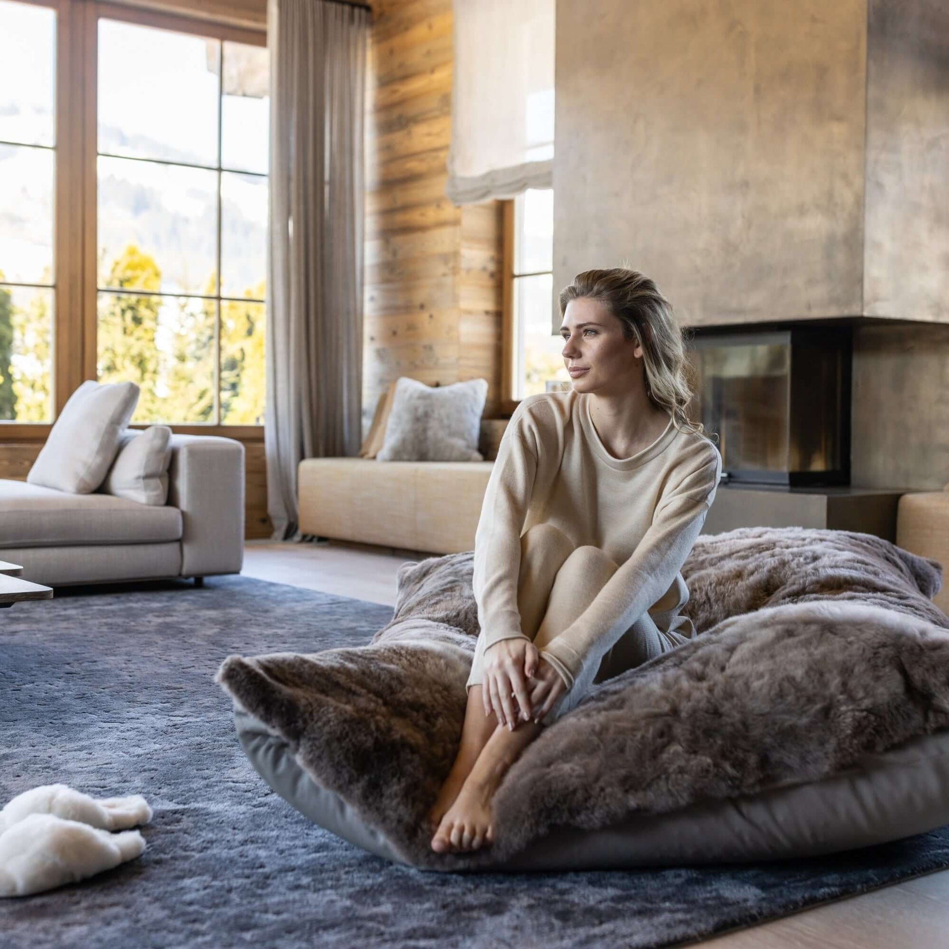Woman sits on Lounger REY, a plush alpaca fur front and leather back lounge, in a cozy living room with large windows.