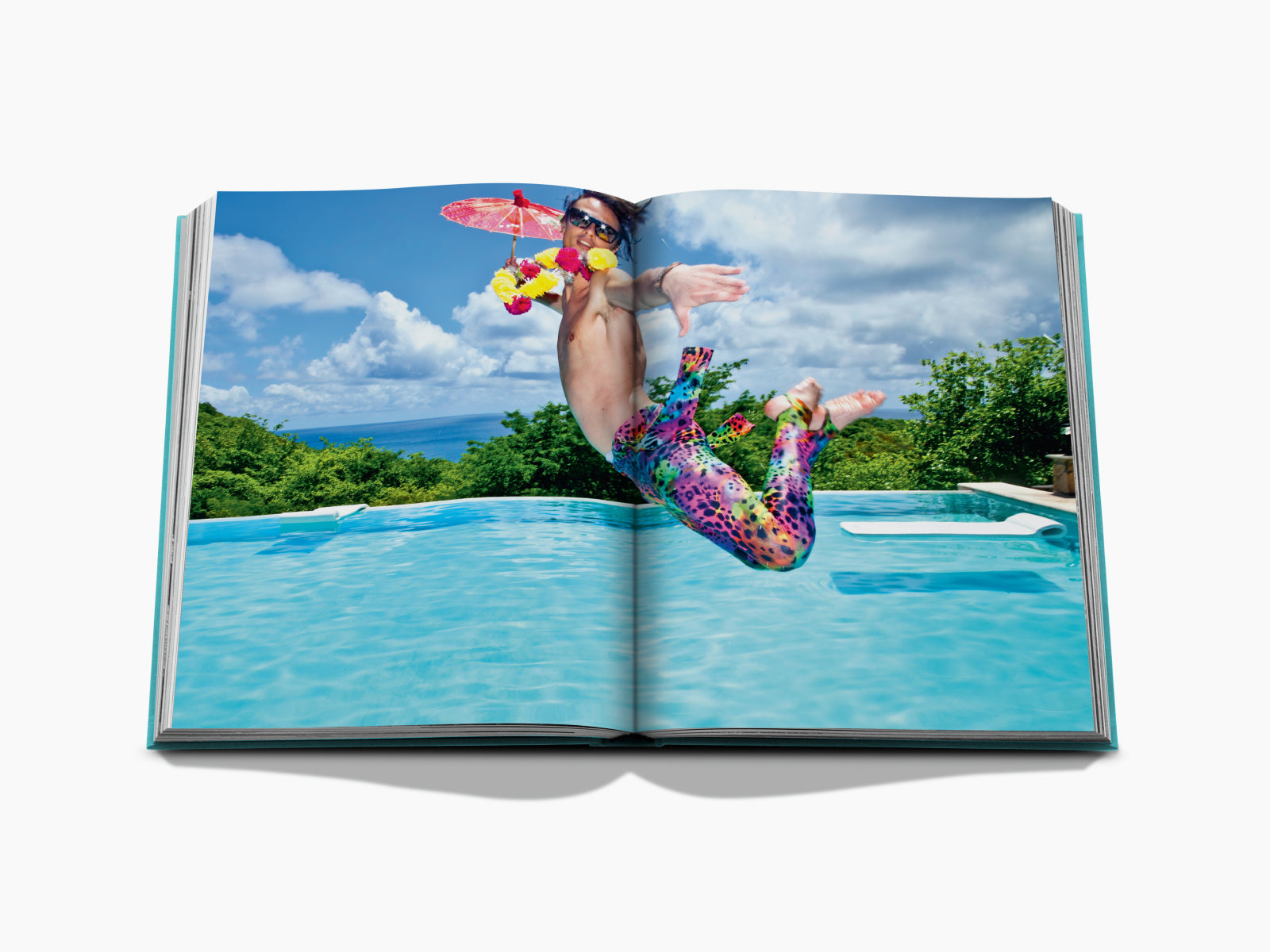 Man in vibrant patterned pants leaps into a pool with a tropical backdrop behind.