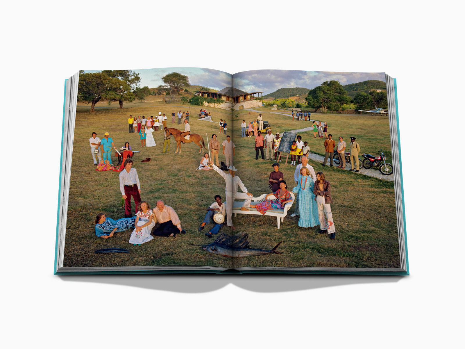 Outdoor lawn gathering with many guests in bright attire, centered around a white bench and a painting on an easel in the background