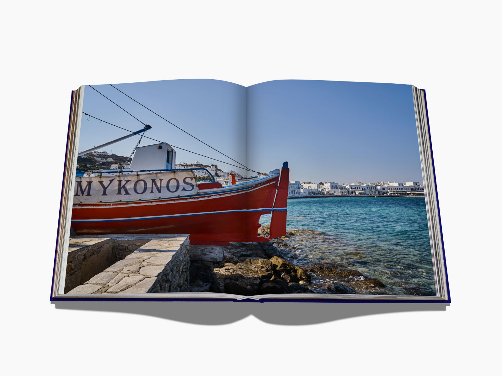 Two page spread of a Mykonos harbor scene with a red boat on rocky shore and whitewashed buildings across turquoise water.