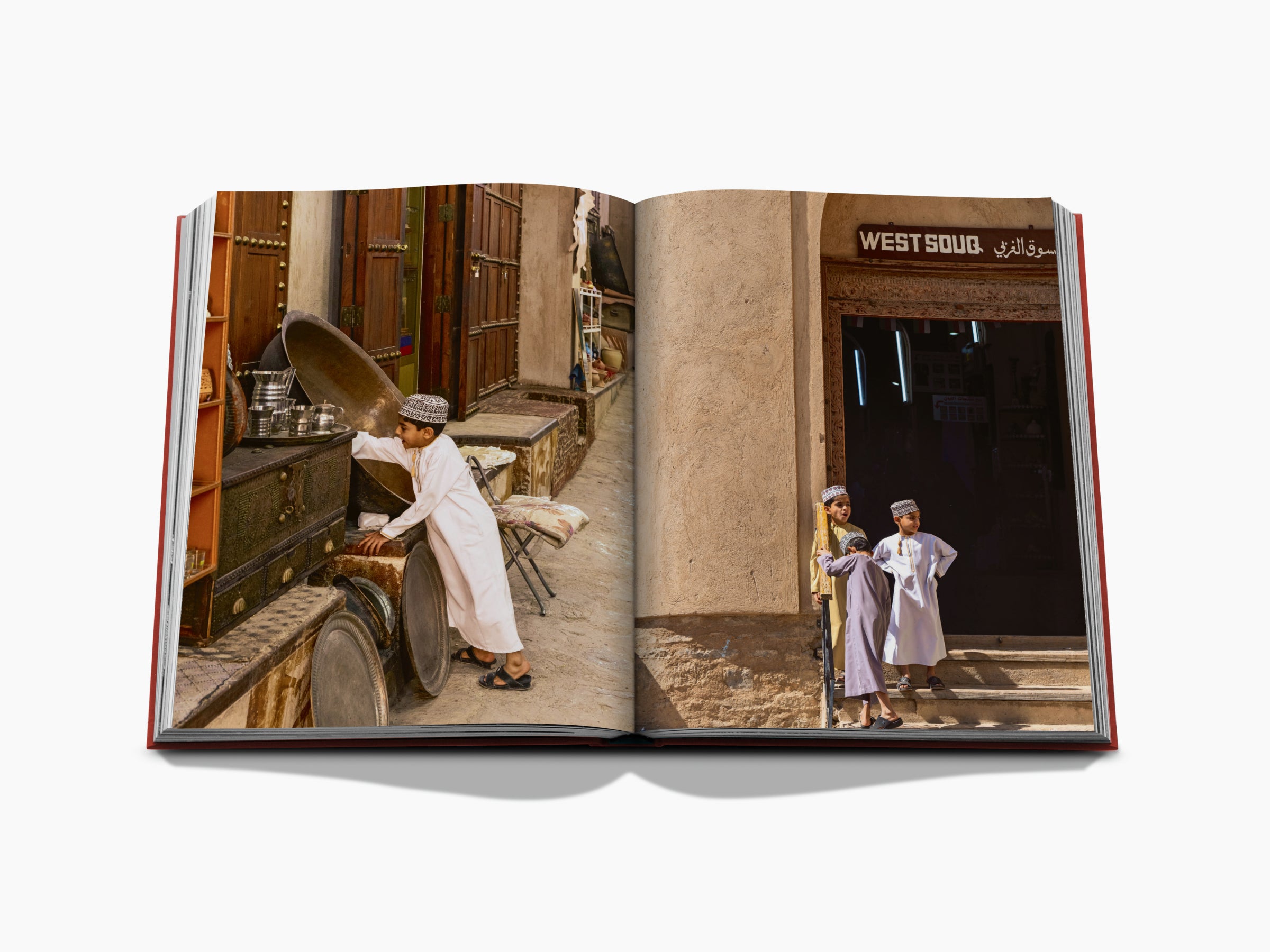 Open travel book spread showing an Omani market scene with a boy cleaning metalware and two children by a doorway.