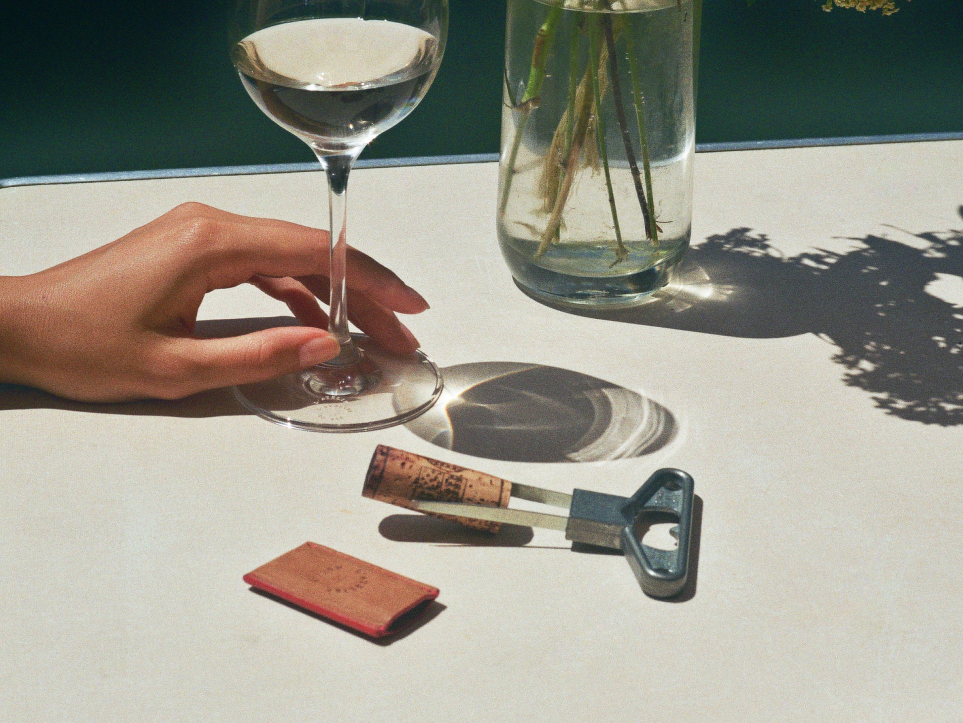 Tabletop scene with a lever corkscrew a cork and a wine glass held by a hand on a pale surface