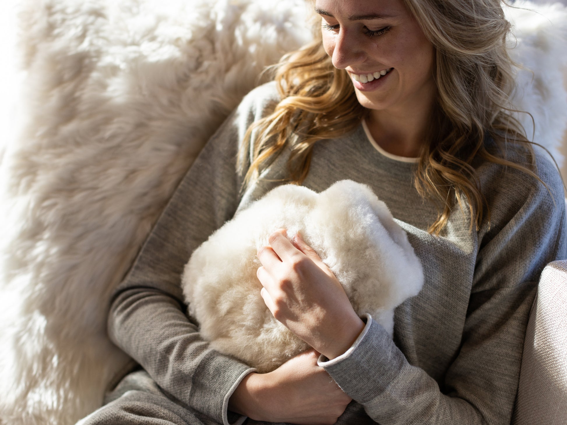 Smiling woman in a grey sweater holding a white fluffy alpaca fiber hot-water bottle cover