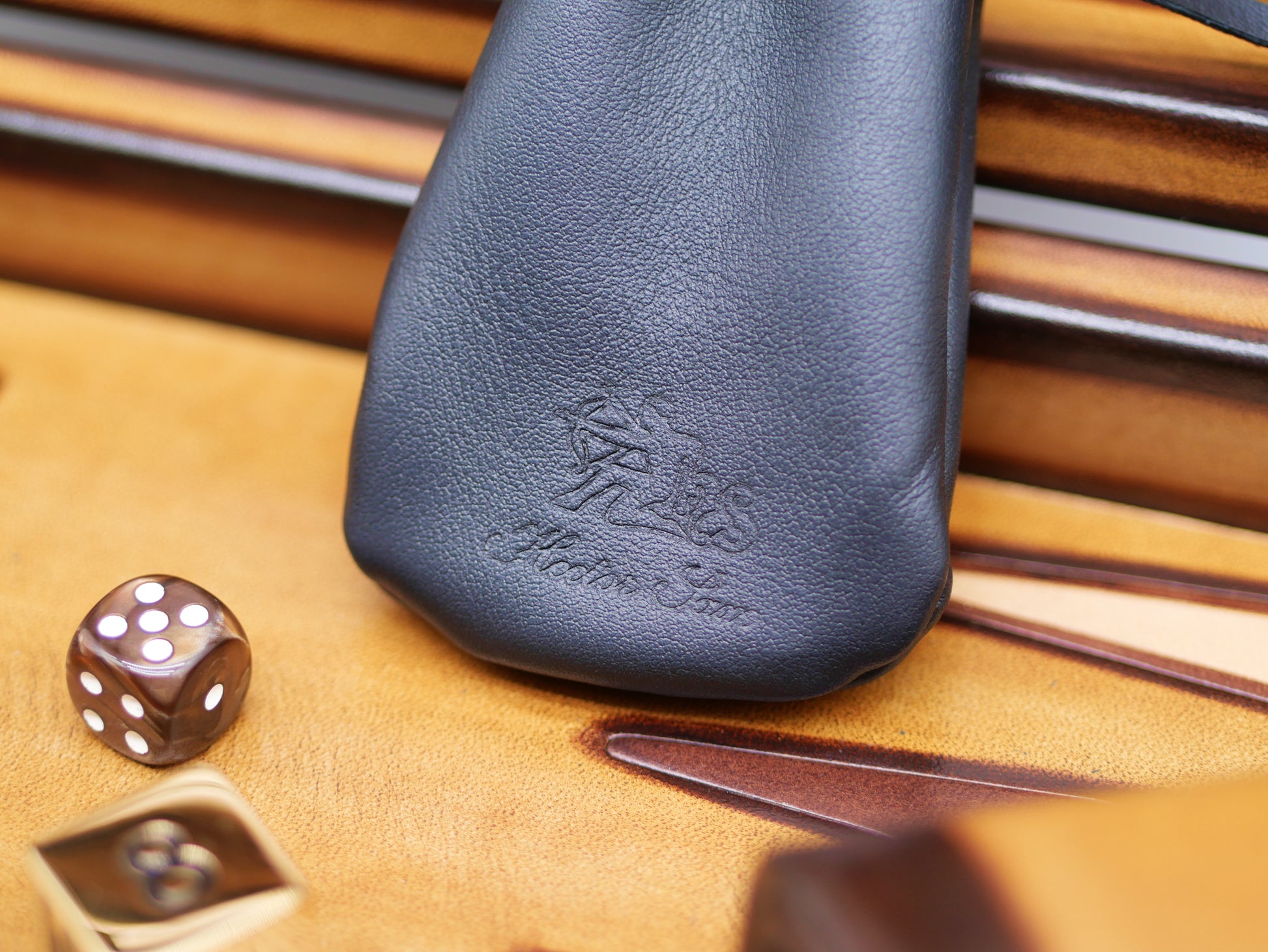 Patinable leather backgammon case with embossed logo sits on a wooden game board near a six-sided die.