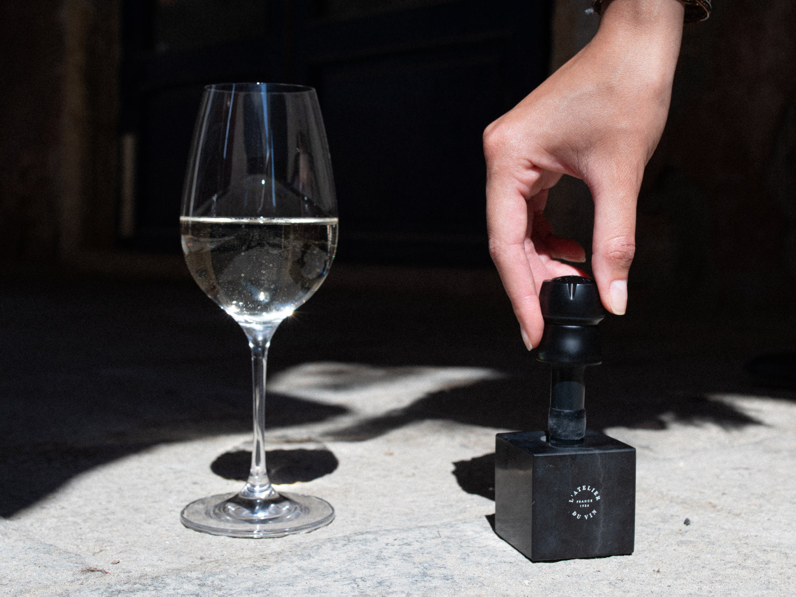 Hand using Bubble Cork Black stopper beside a wine glass on a sunlit surface.