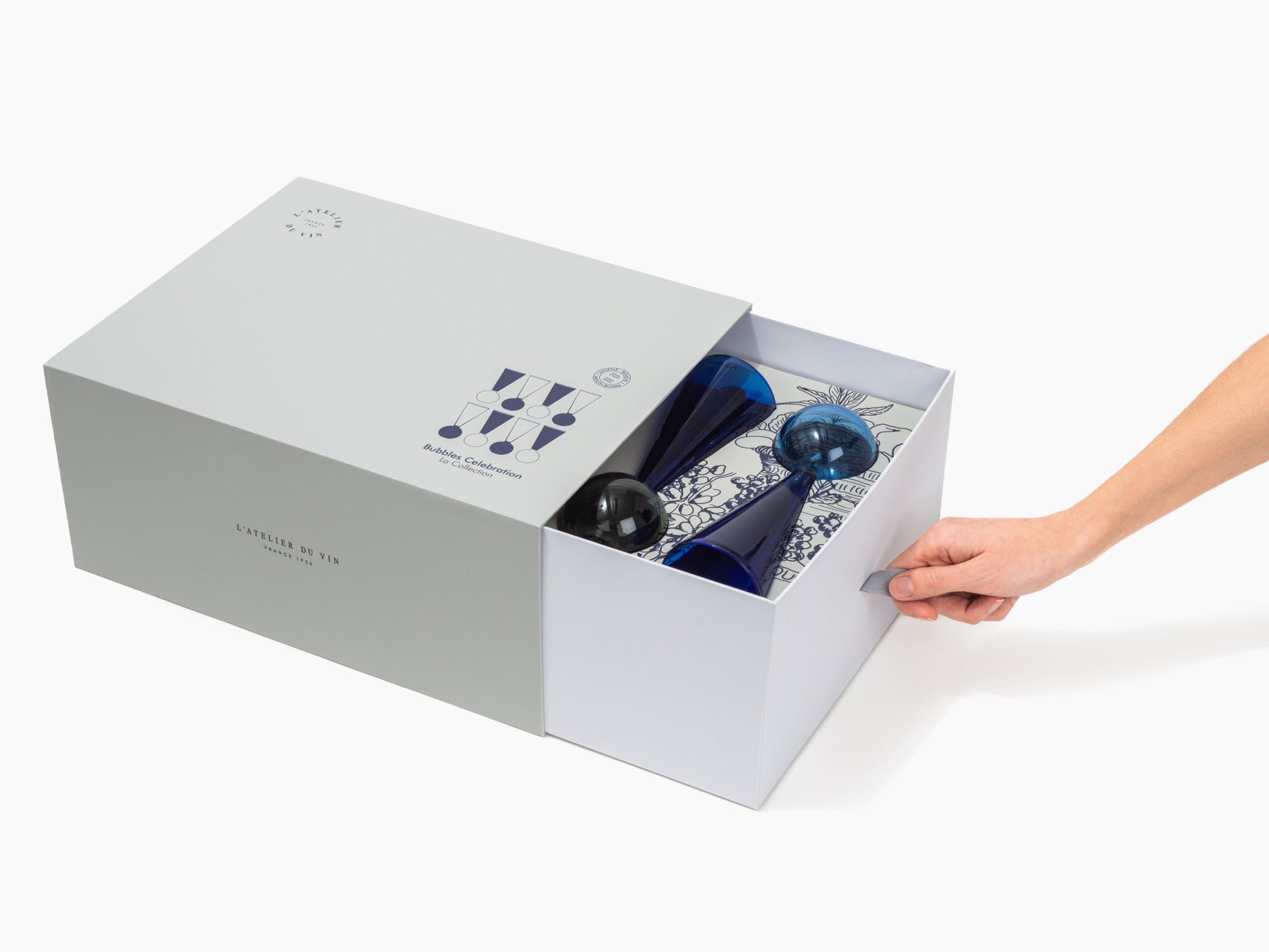 Open Bubbles Celebration box revealing blue glassware and accessories in a decorative insert as a hand pulls the tab