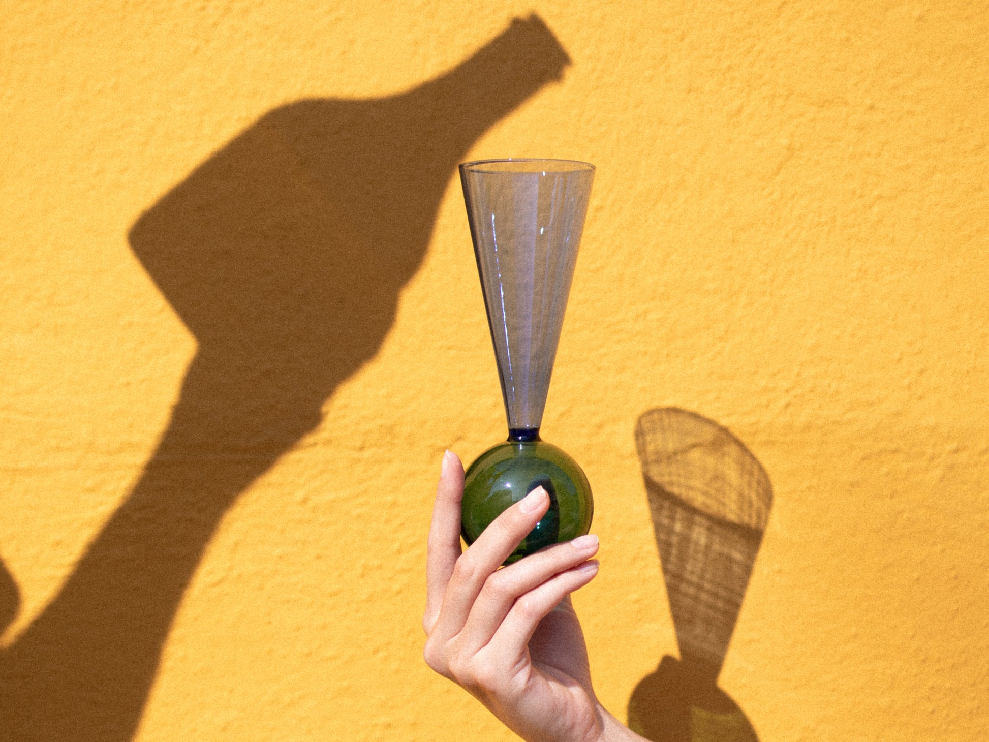 A hand holds a blue tapered Bubbles Celebration flute with a green bulbous base against a bright yellow wall and strong shadow