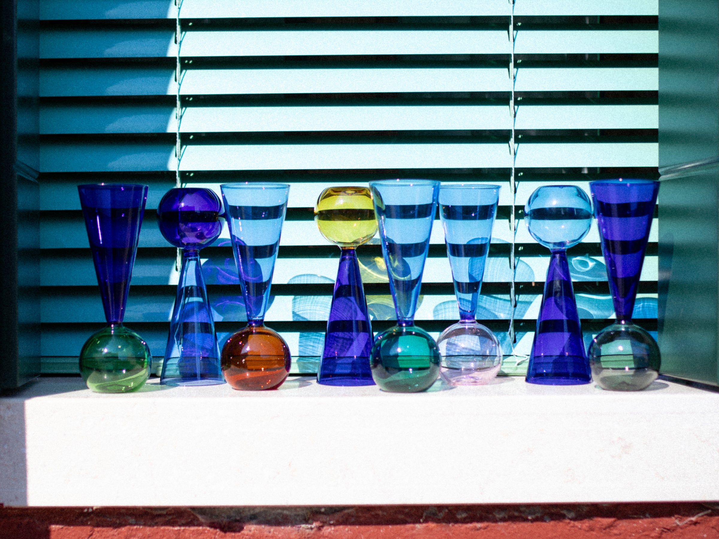 Eight Bubbles Celebration flutes in blue and multicolored glass arranged in a row on a white ledge with a striped window backdrop