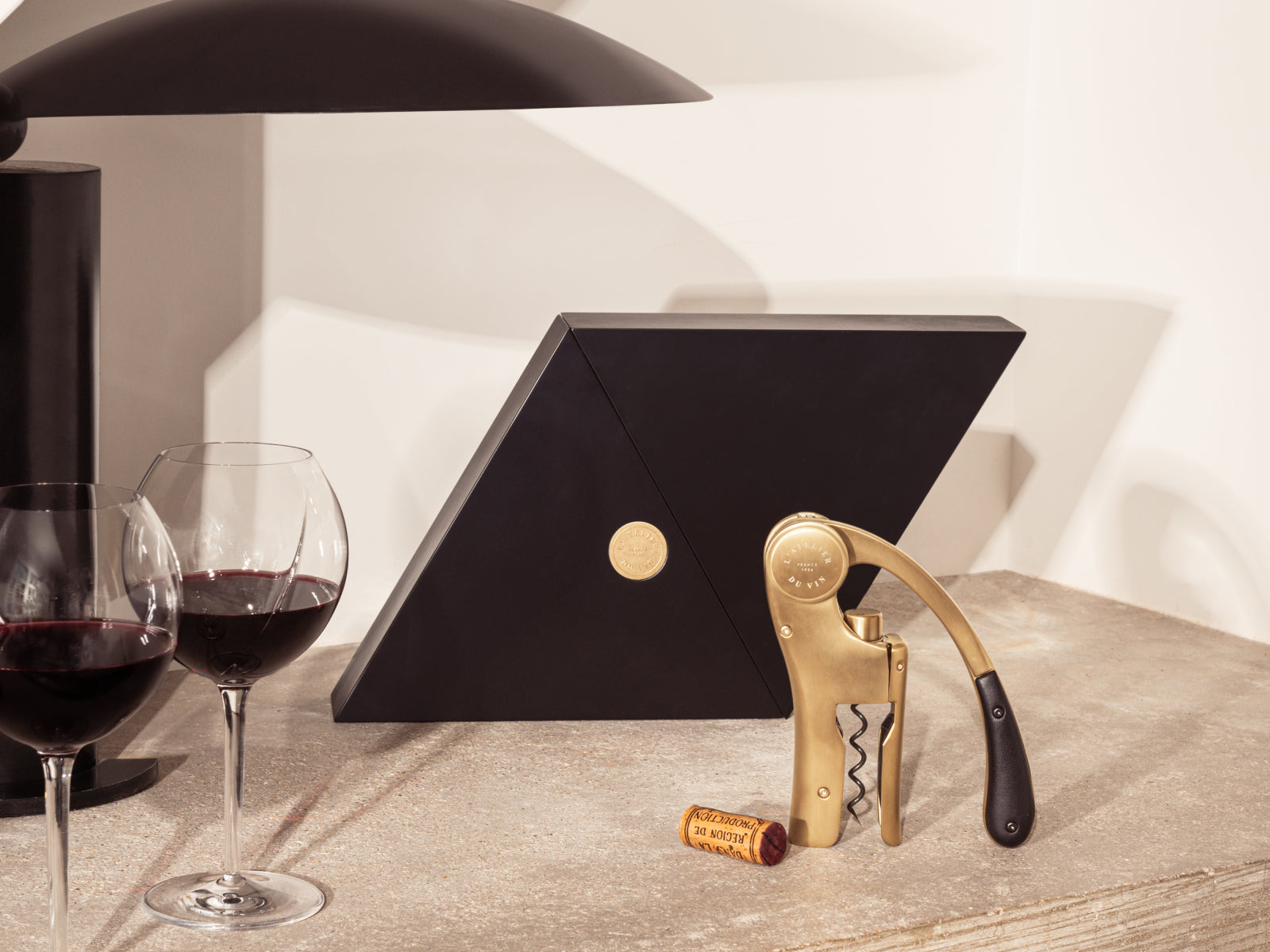 Geometry black lacquered wood box opens to reveal the gold Oeno Motion corkscrew on a neutral countertop beside wine glasses