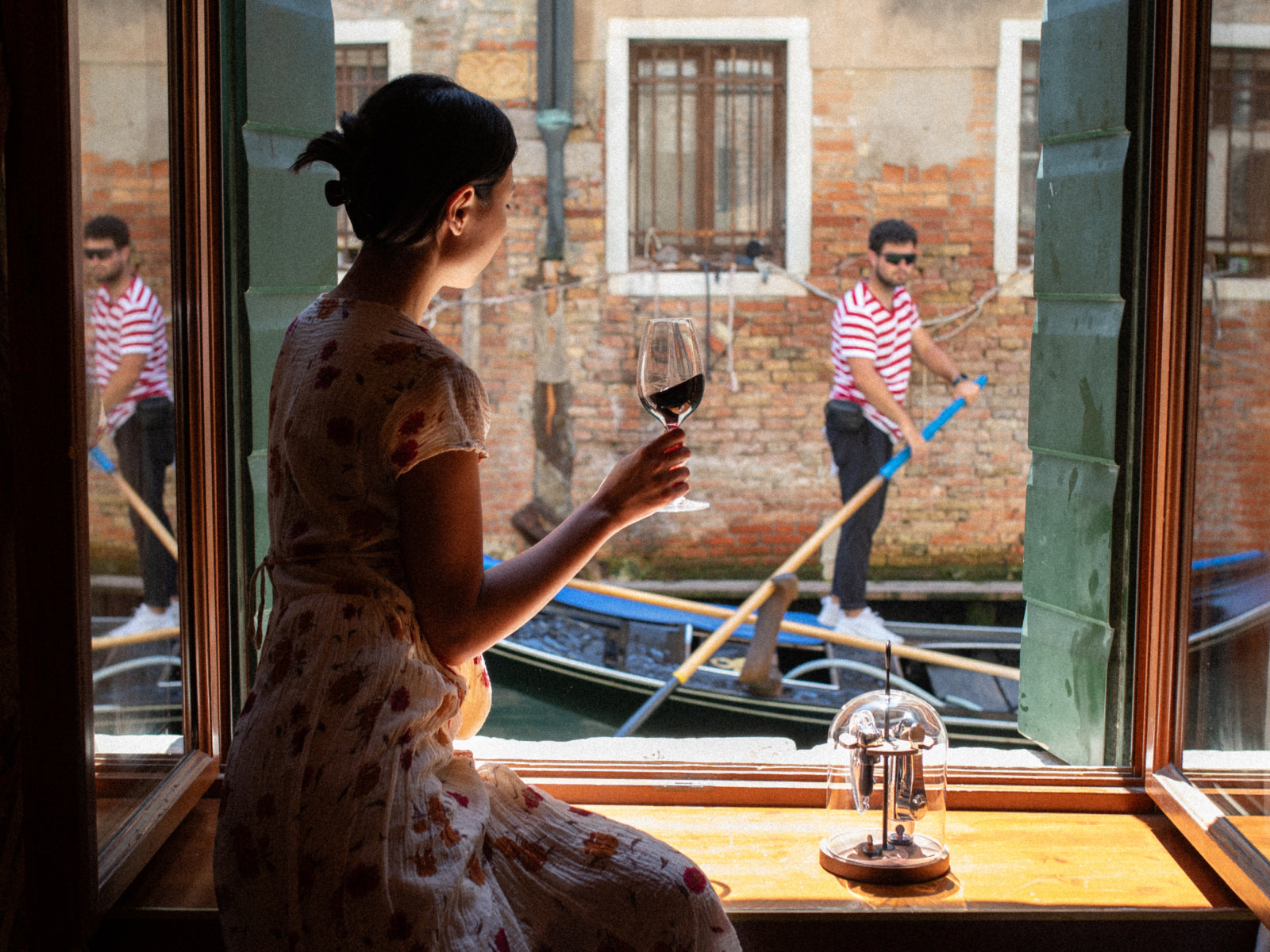 Woman in a floral dress gazes from a Venetian window holding a wine glass with a Le Globe on the sill