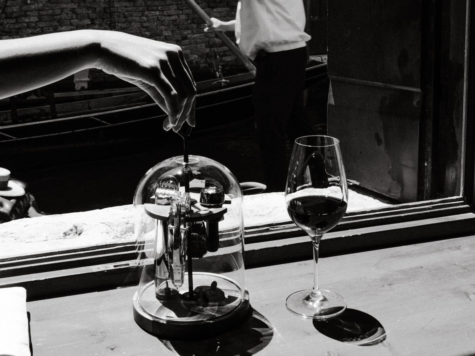 Hand adjusts a corkscrew under a glass dome on a walnut base beside a glass of red wine