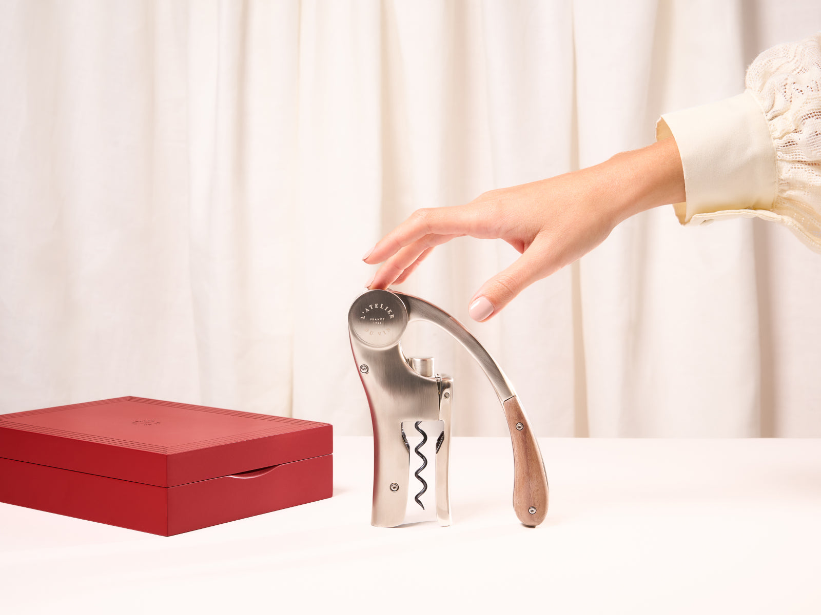 Hand reaching toward a curved stainless steel lever corkscrew beside a red lacquered engraved gift box.