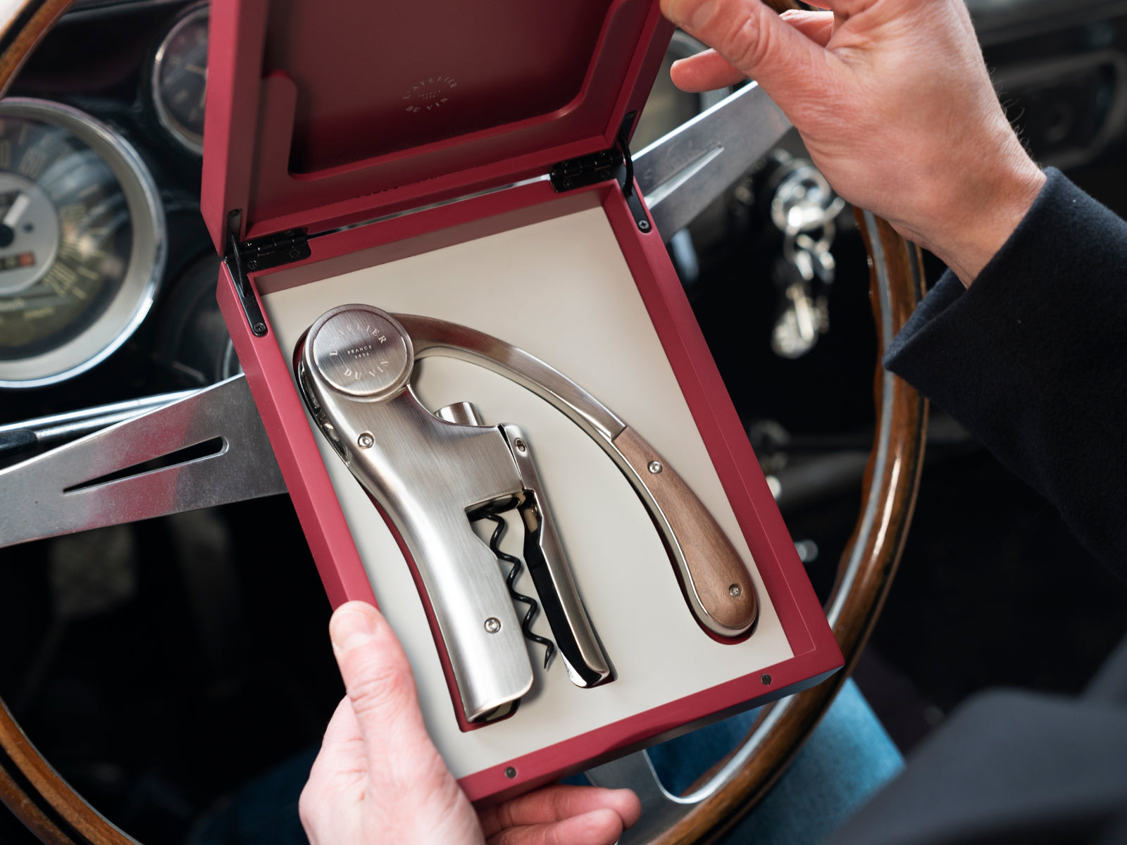 Hands hold an open red oenology case in a car interior, revealing the lever corkscrew with wooden handle.