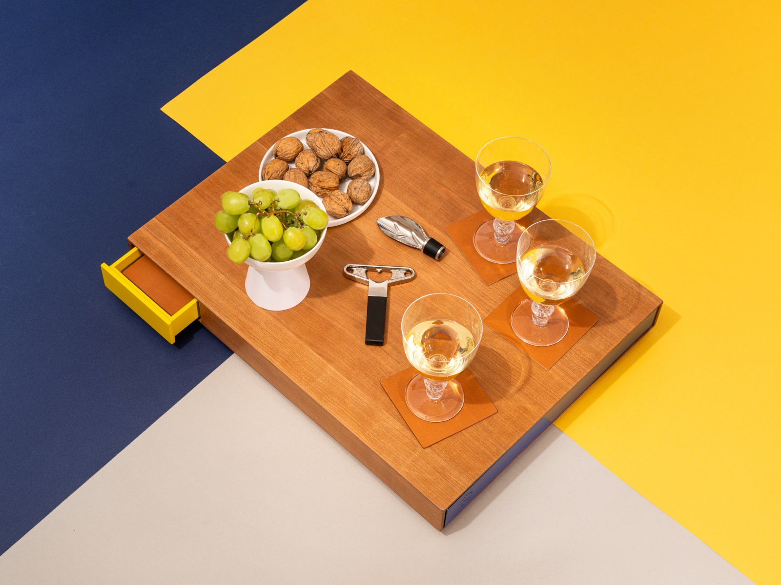 Wooden tasting board on a bold blue and yellow geometric backdrop with three white wine glasses, bowls of grapes and walnuts, and a corkscrew.
