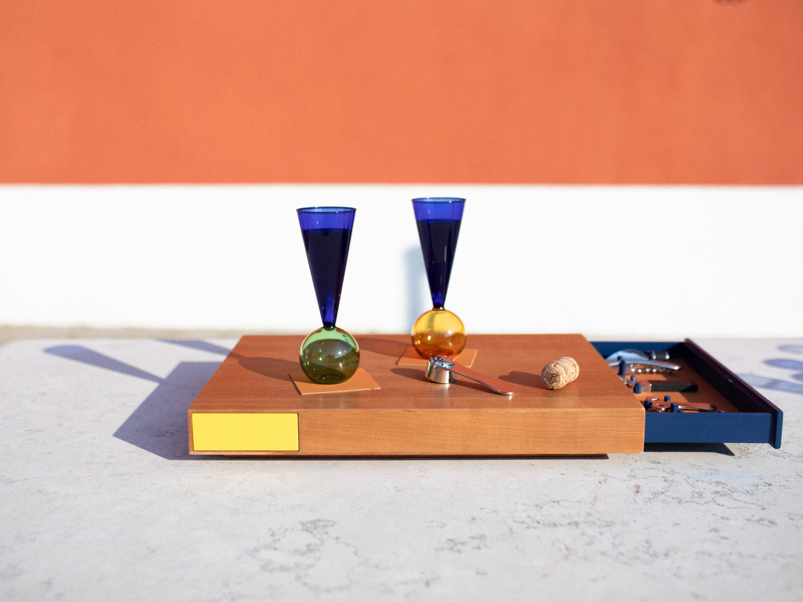 Two cobalt blue stem glasses with colored spherical bases rest on a wooden board beside a corkscrew and cork.