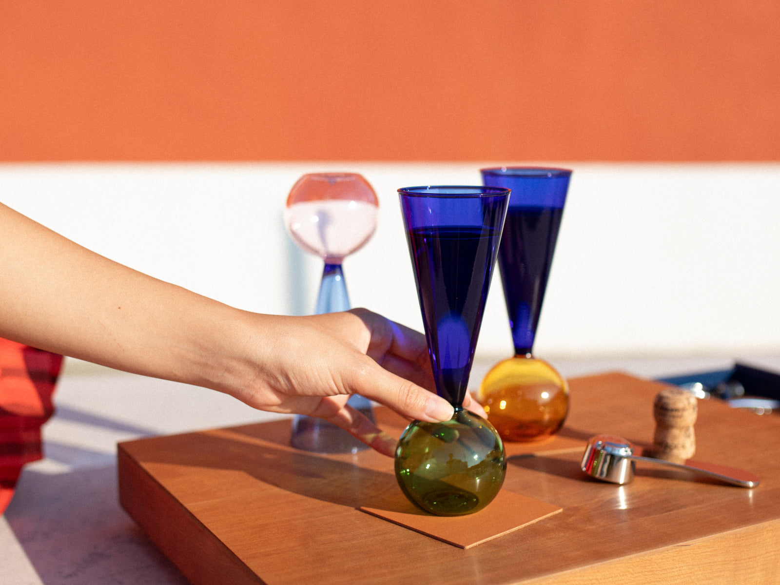 Hand places a green glass base on a wooden Le Plateau serving board with cobalt blue stemmed glasses nearby.