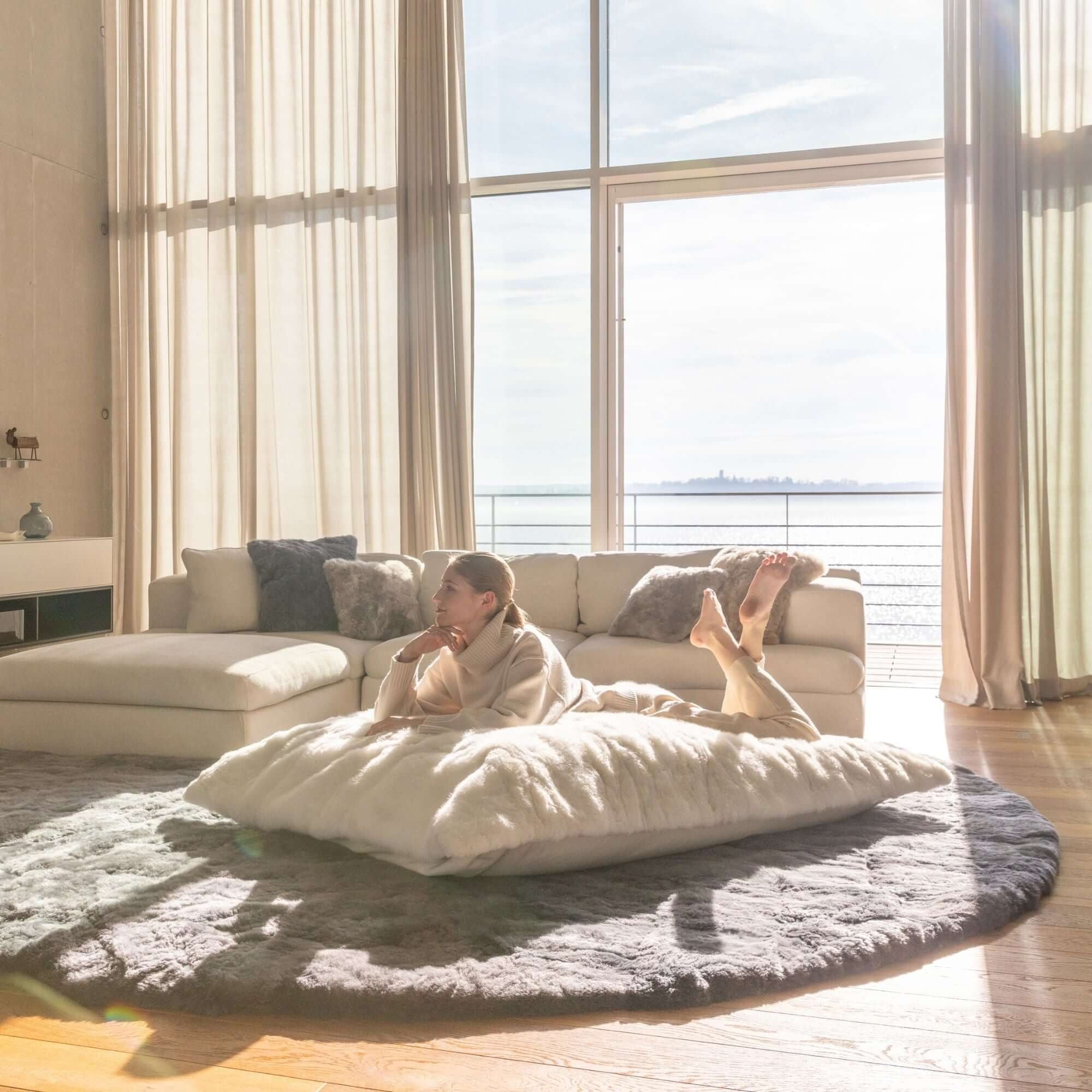 Person lounging on Lounger REY made of white alpaca fur in a sunlit living room with a round rug and beige sofa.