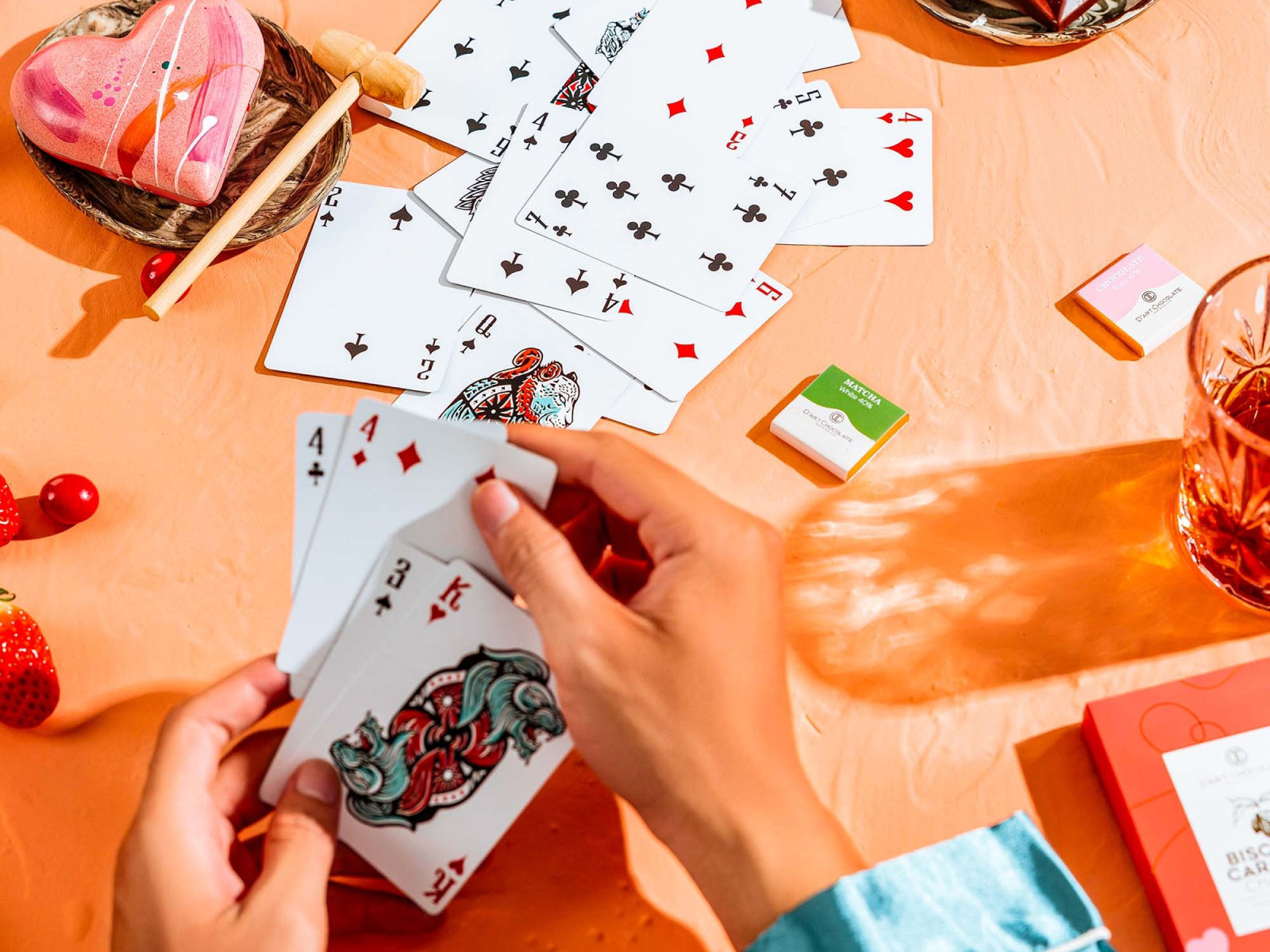 Hands hold a card among scattered Animal Kingdom playing cards on a peach table with colorful chocolate boxes and glasses nearby.
