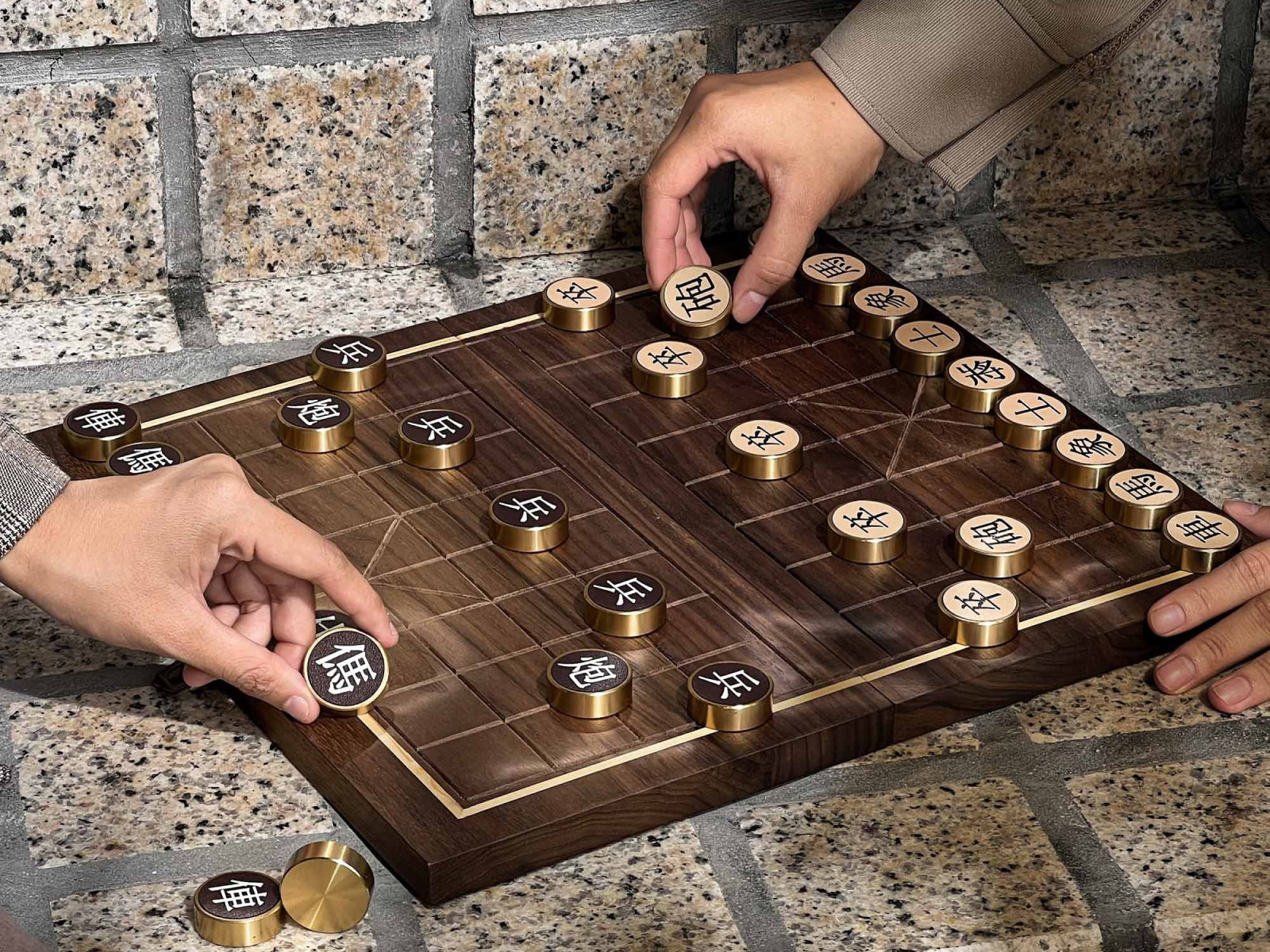Two players move bronze Chinese chess pieces on a walnut board set on a stone surface outdoors.