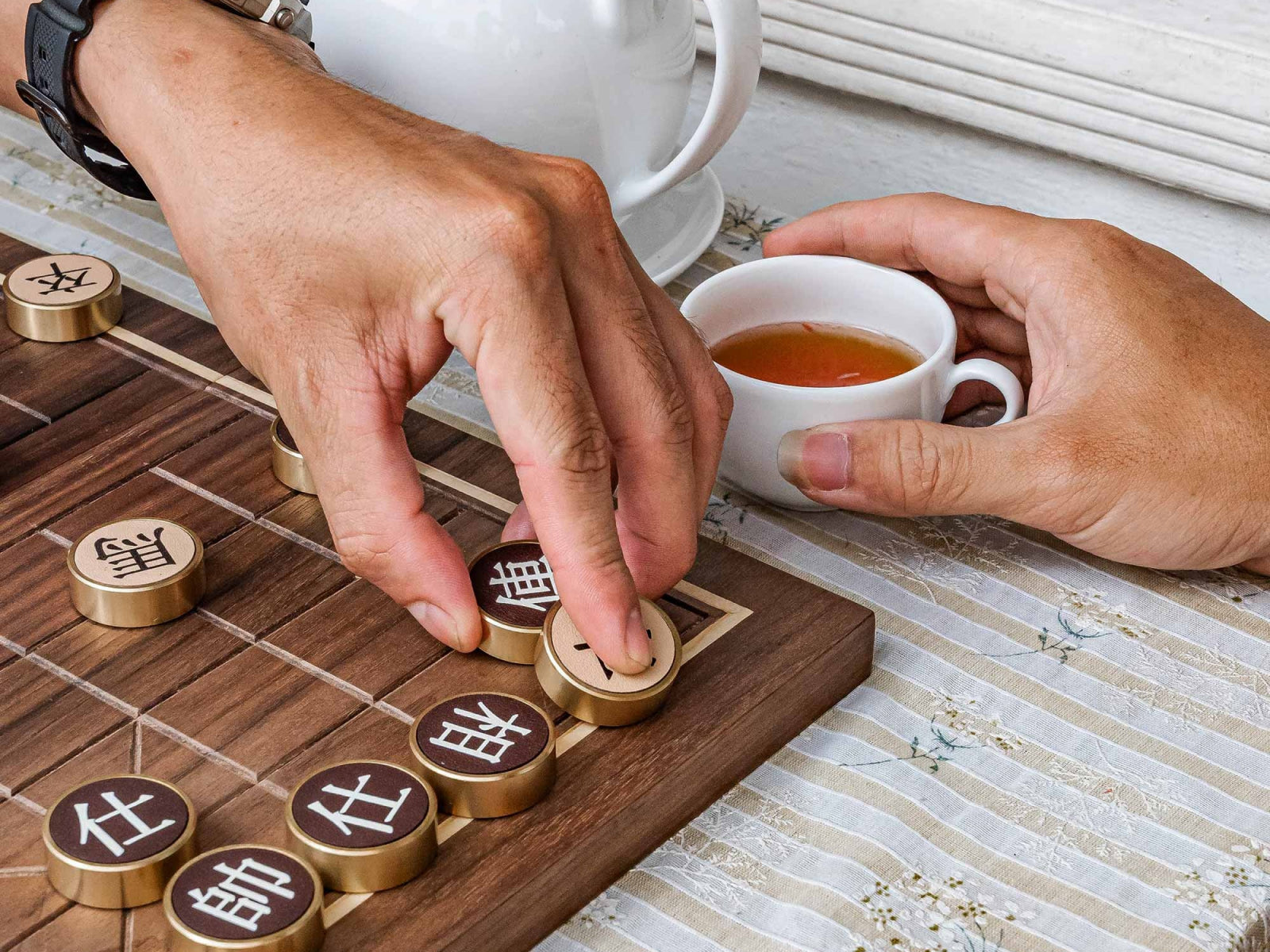 Hands place bronze Chinese chess pieces on a walnut Compact Chinese Chess board beside a teacup.
