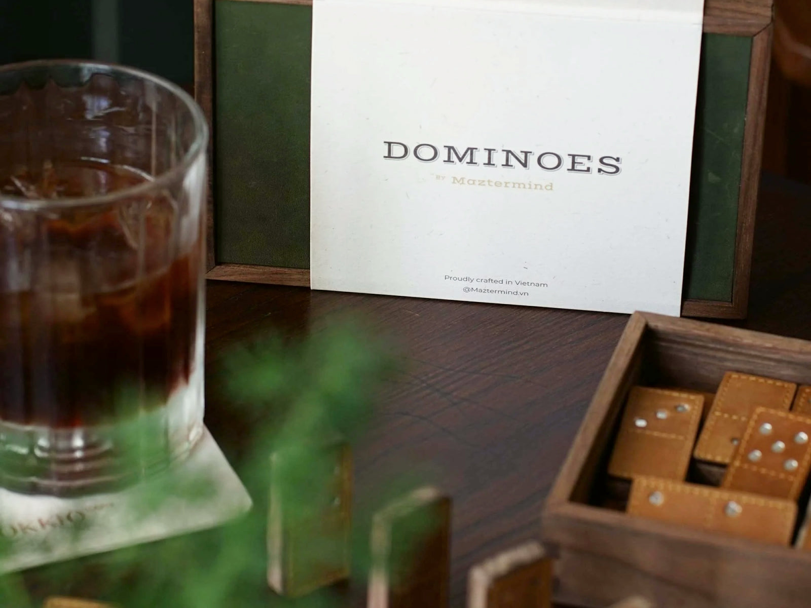Leather Dominoes Set packaging with a white DOMINOES card in a wooden frame, beside a glass and a tray of leather domino tiles.