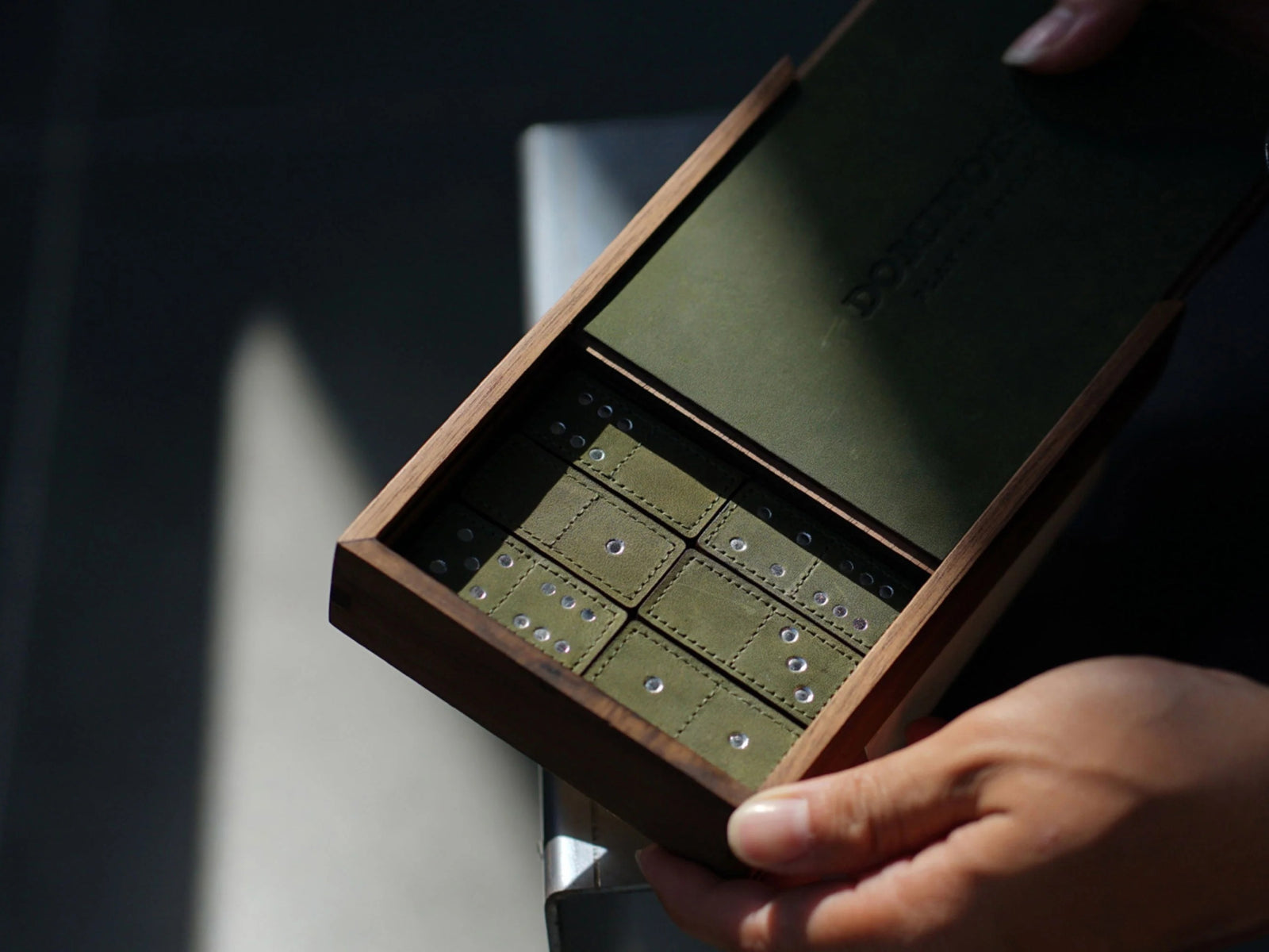 Open wooden box with green interior holding leather domino tiles with metal pips, partially captured by a hand.
