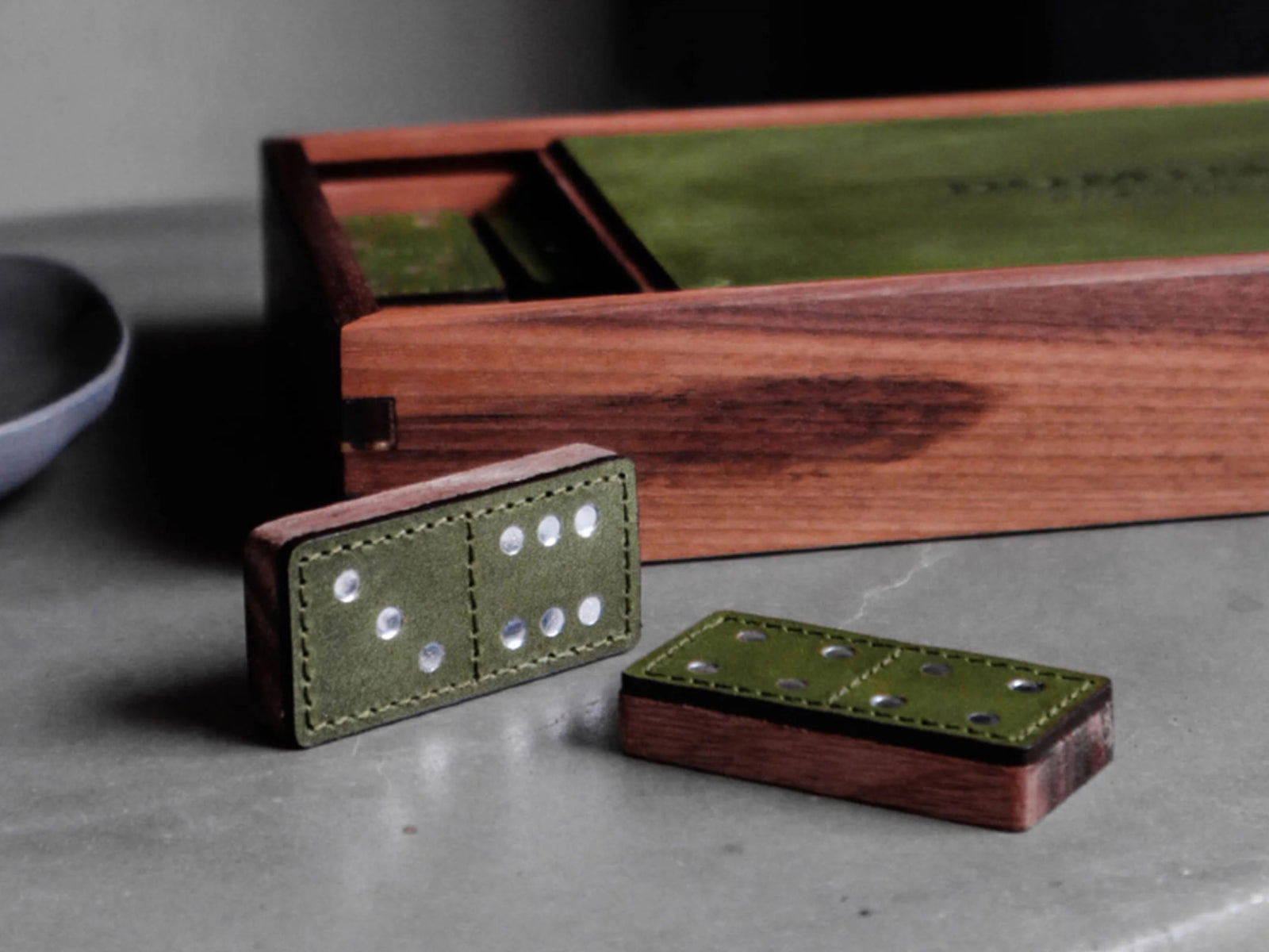 Green leather domino tiles with white pips spread on a wooden table beside a wooden box