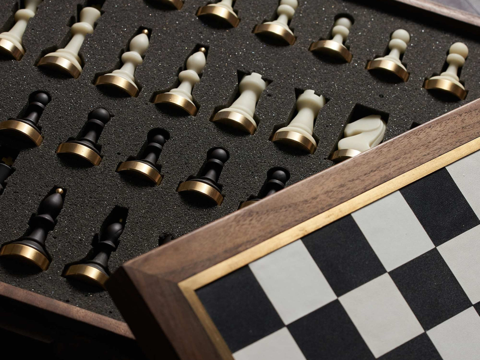Close up of black and ivory chess pieces with brass bases nestled in a foam tray beside a checkered panel