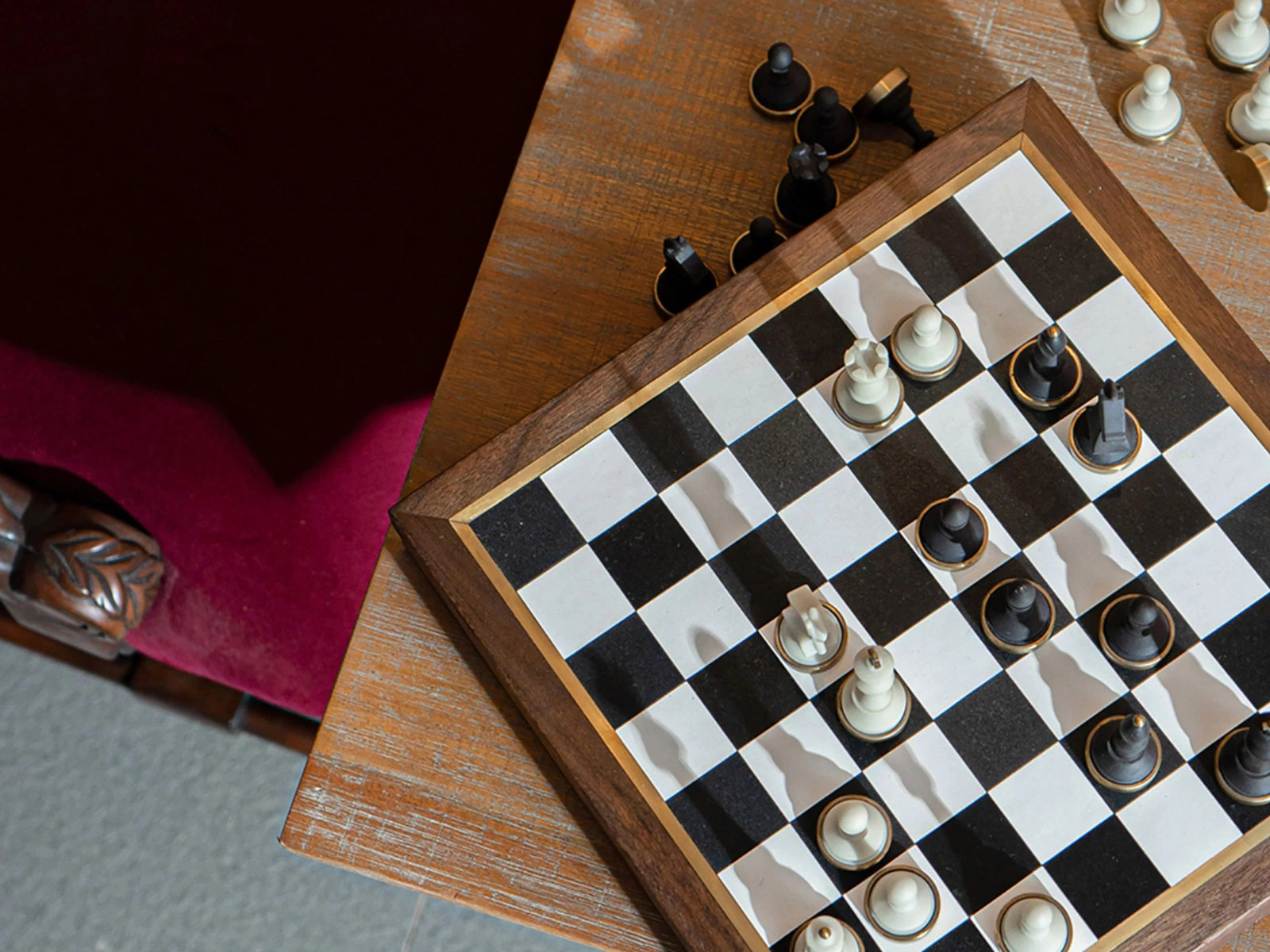 Birds-eye view of a luxury walnut chessboard with white and black pieces arranged for a midgame on a wooden table.