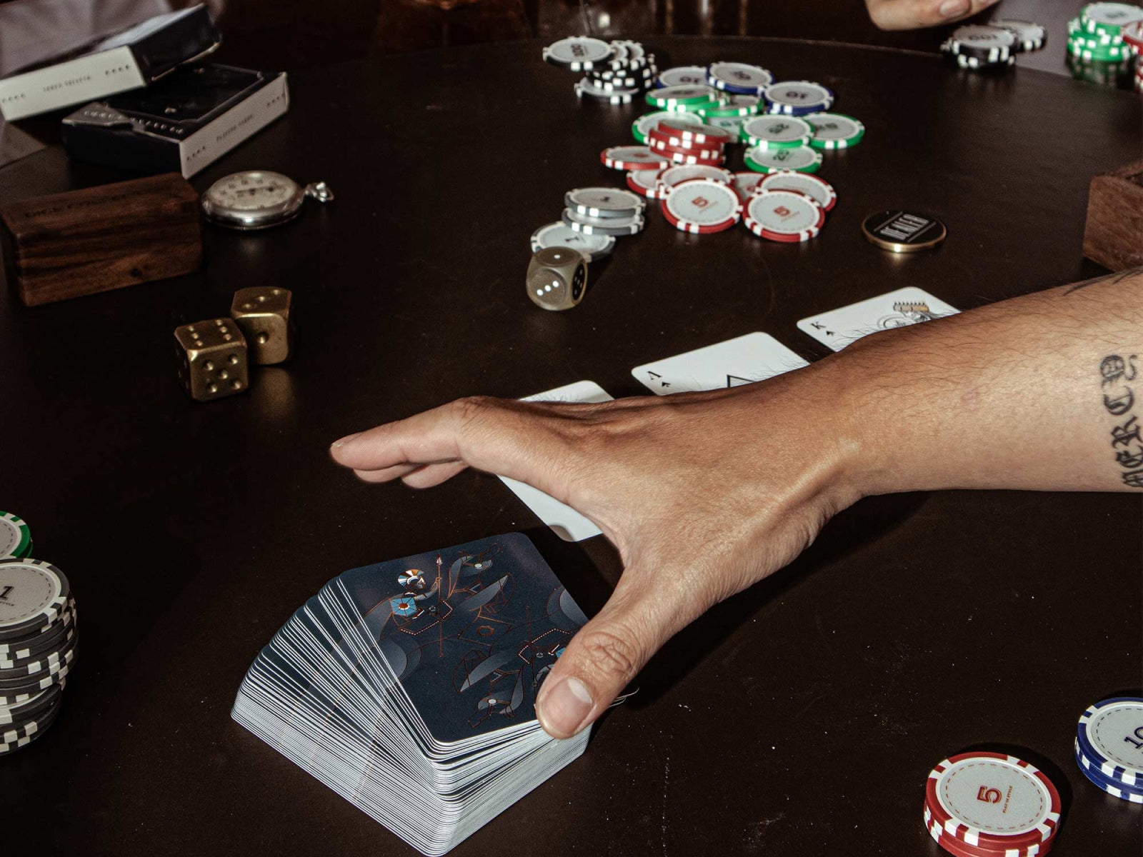 Hand reaches toward a Bowtie Poker Chip Set on a dark table with two premium card decks and stacks of chips.