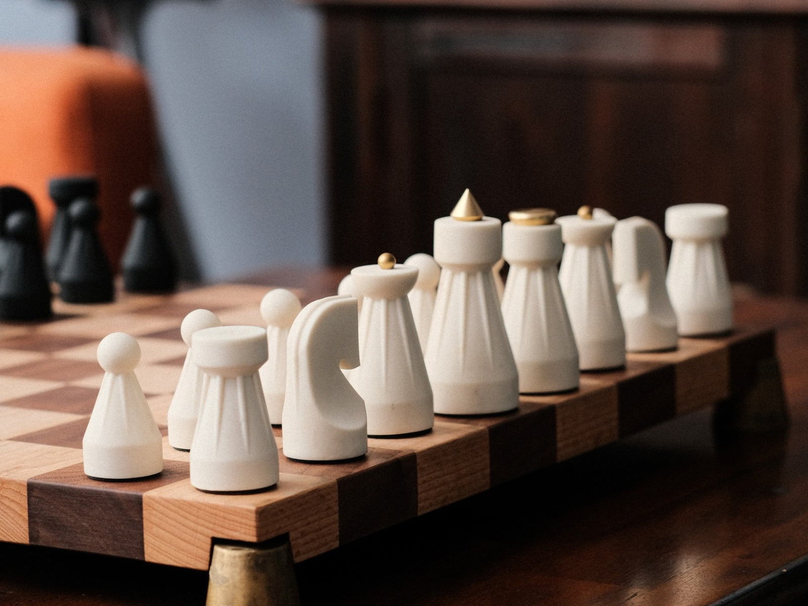 White chess pieces lined up on a wooden checkered chessboard with blurred black pieces in the background