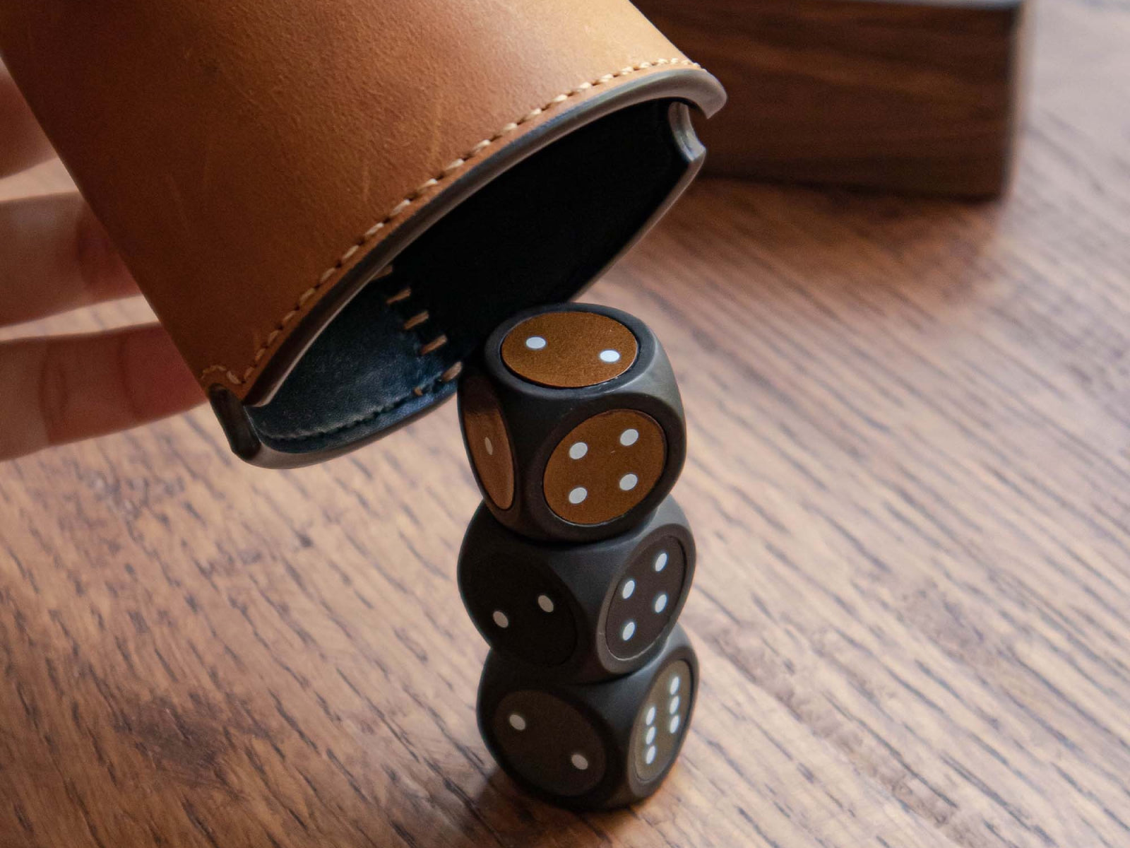 Leather wrapped dice cup with tan stitching on a wood surface, brown dice with white pips stacked beside it.