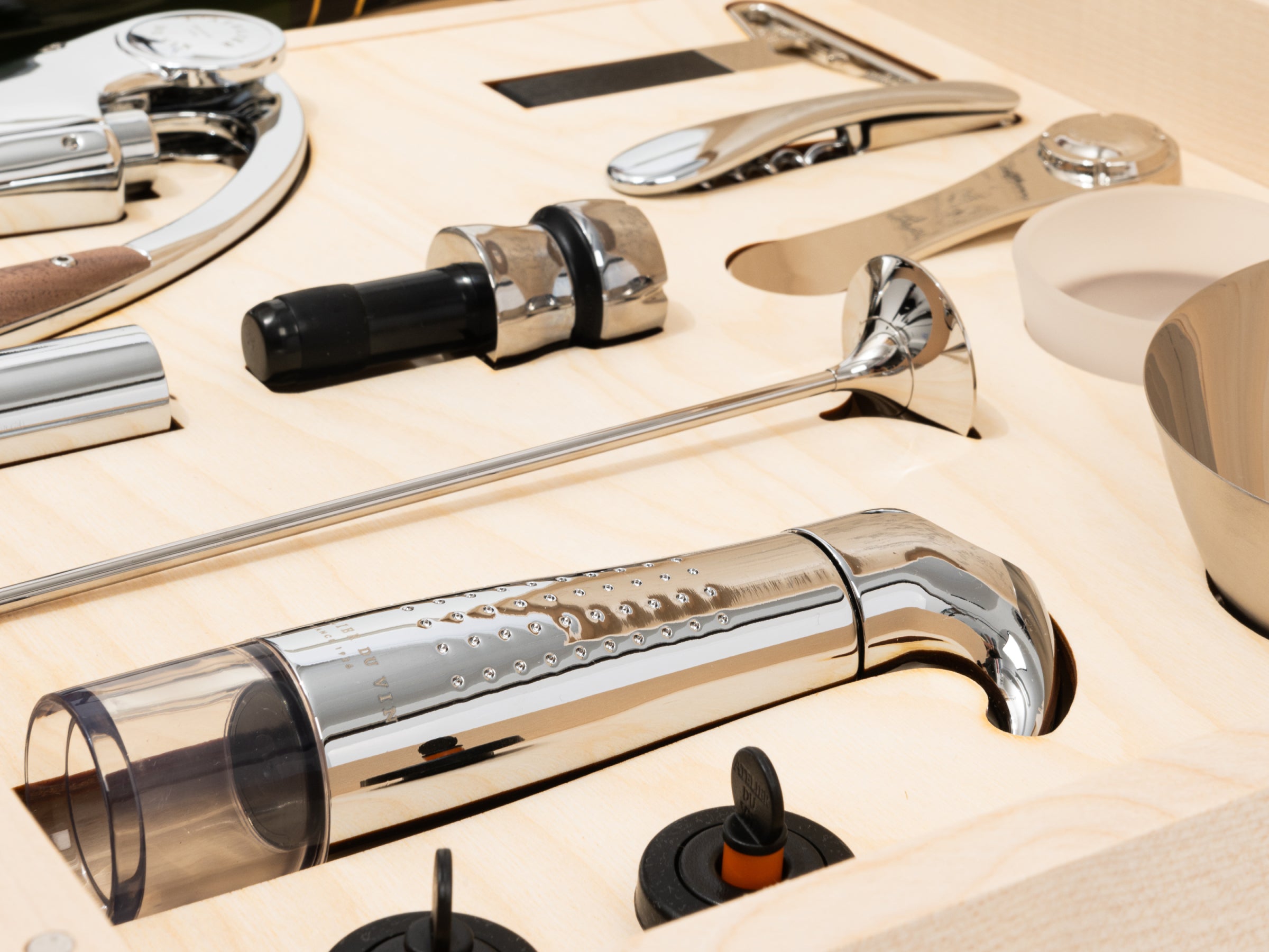 Polished wine tools including corkscrew and stopper laid out on a light wood surface beside a wooden case.