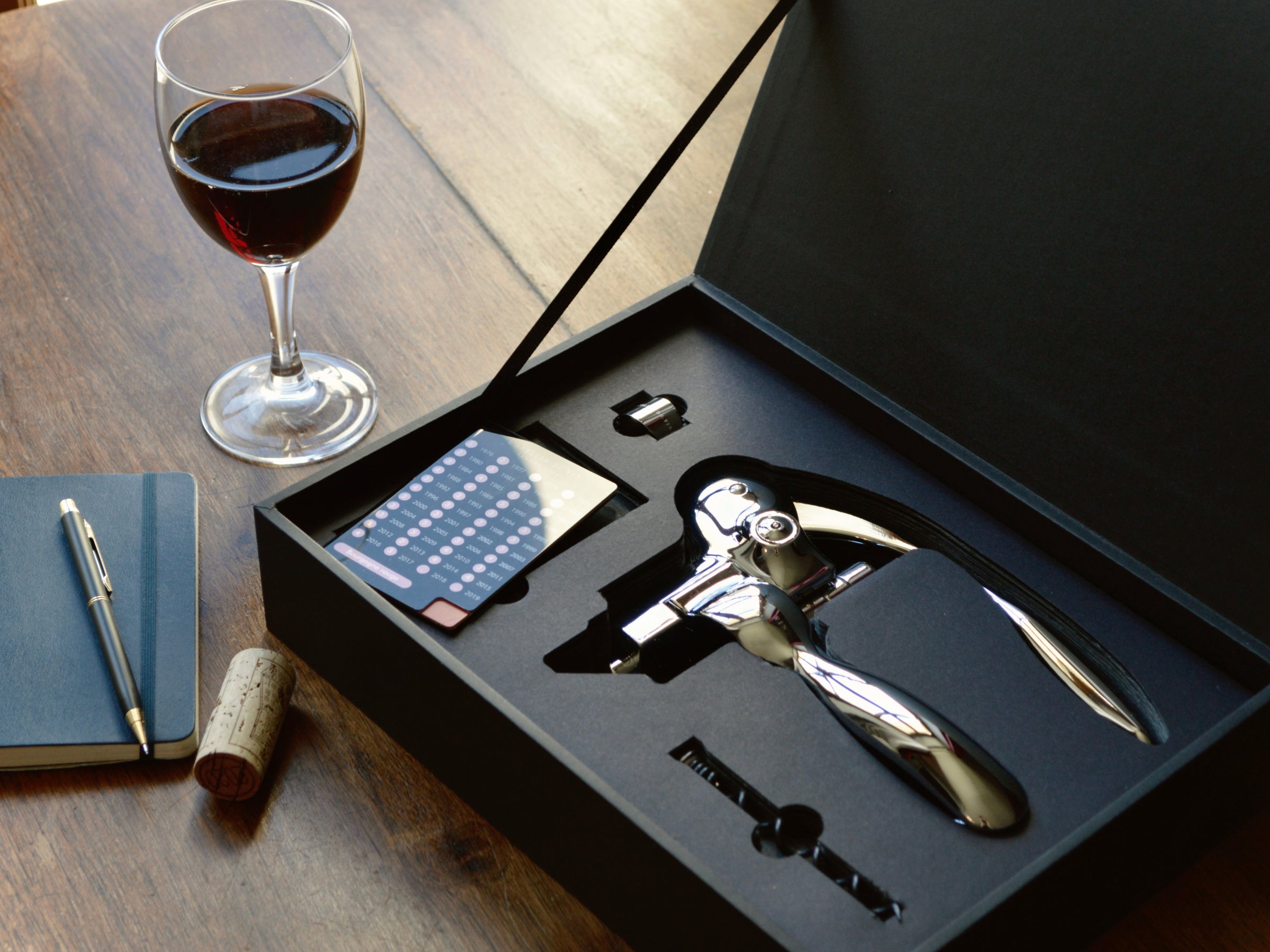 Open black gift box displaying a chrome lever corkscrew and accessories on a wooden table with a glass of red wine nearby