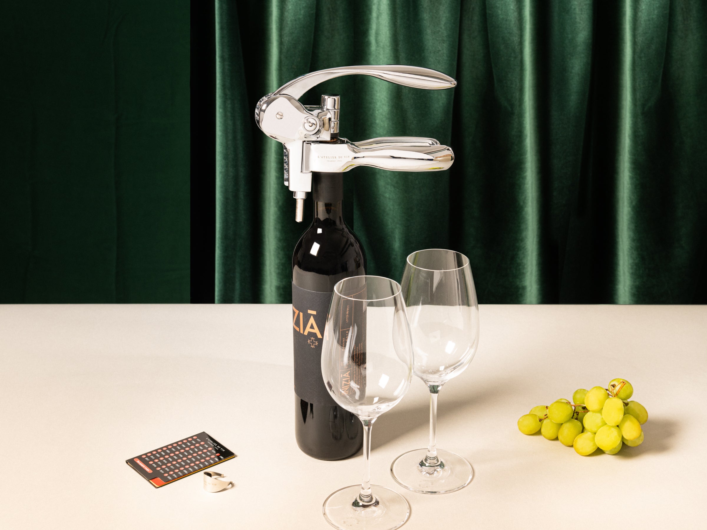 Lever corkscrew perched on a wine bottle with two stemmed glasses and a grape cluster against a green velvet backdrop.