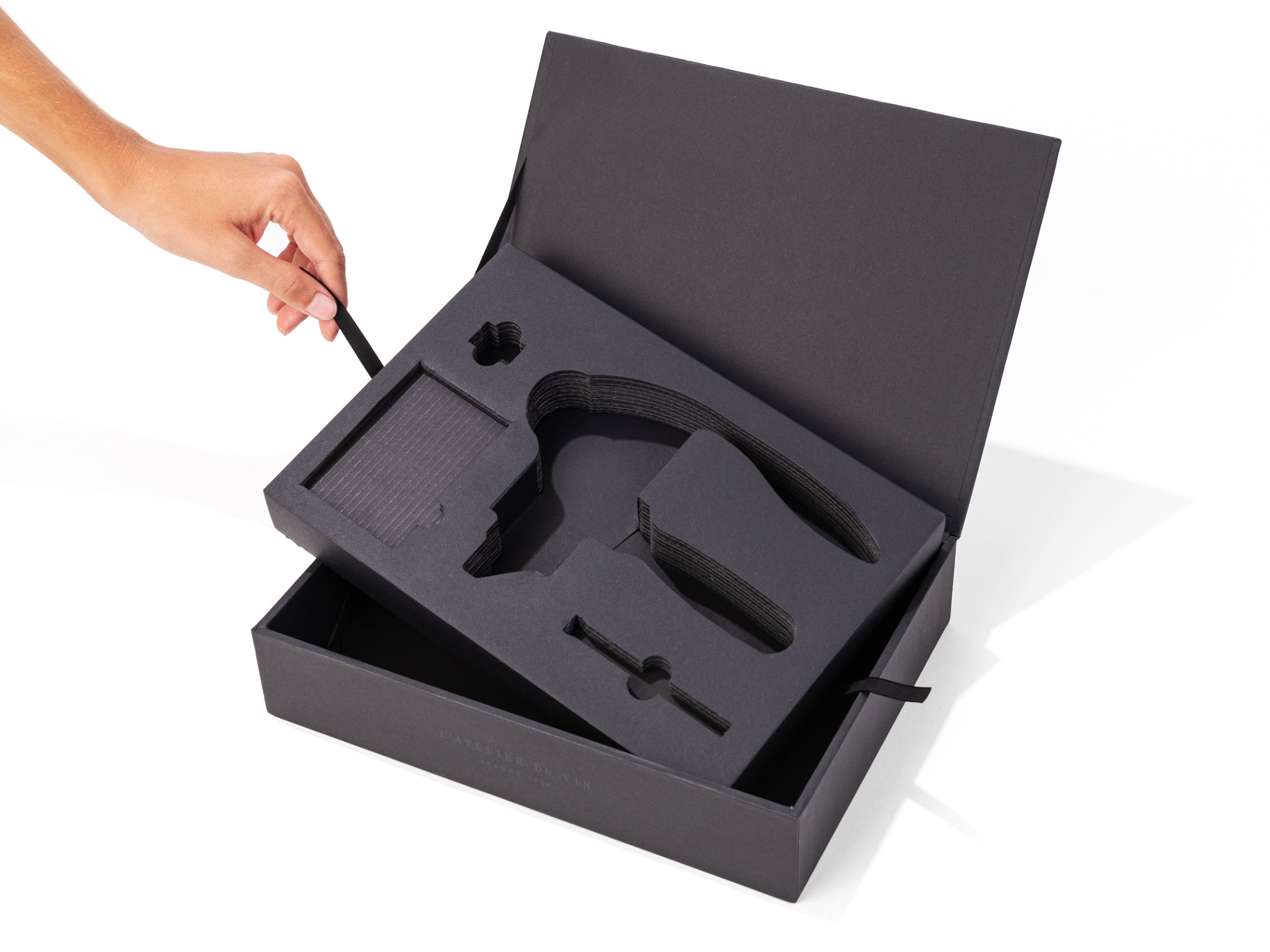 Opened dark presentation box for Oeno Box Sommelier revealing foam insert and tool slots with a hand lifting a ribbon.