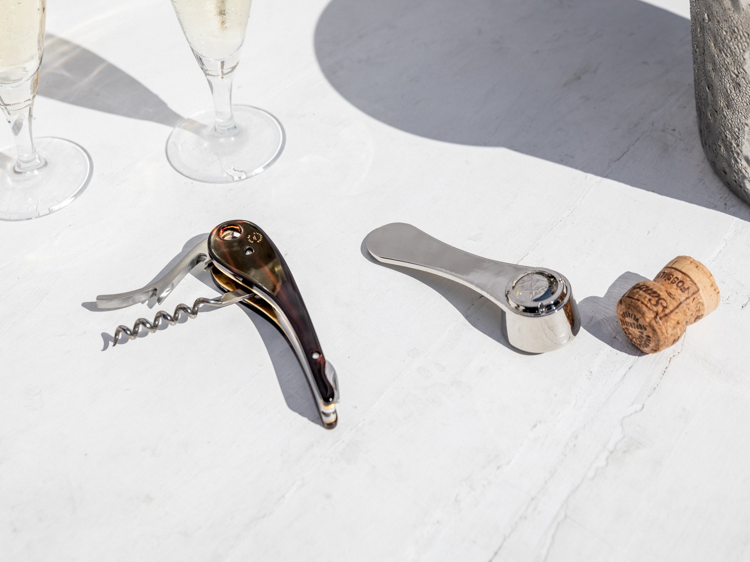 Wood handled corkscrew and silver lever opener laid on a white surface with a cork nearby outdoors