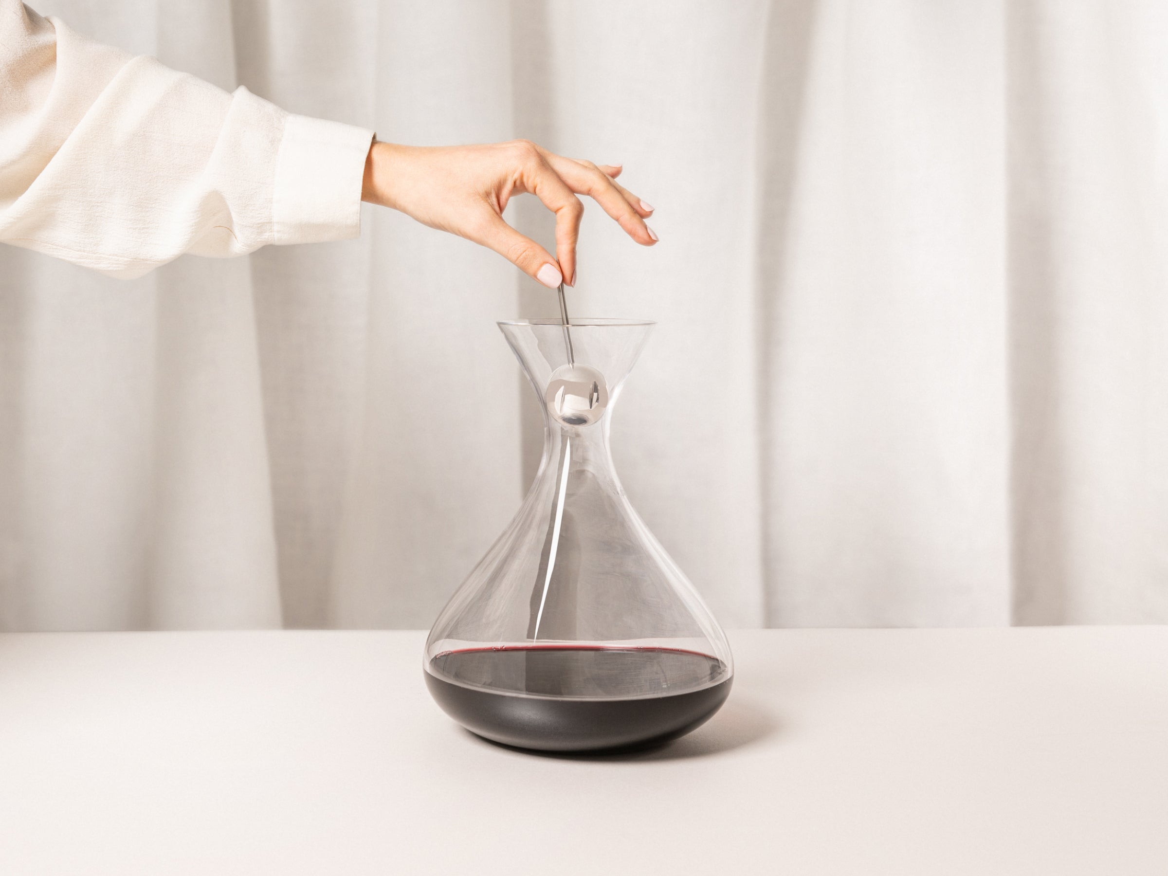 Hand in a white sleeve inserts a metal wand into a clear glass decanter containing red wine