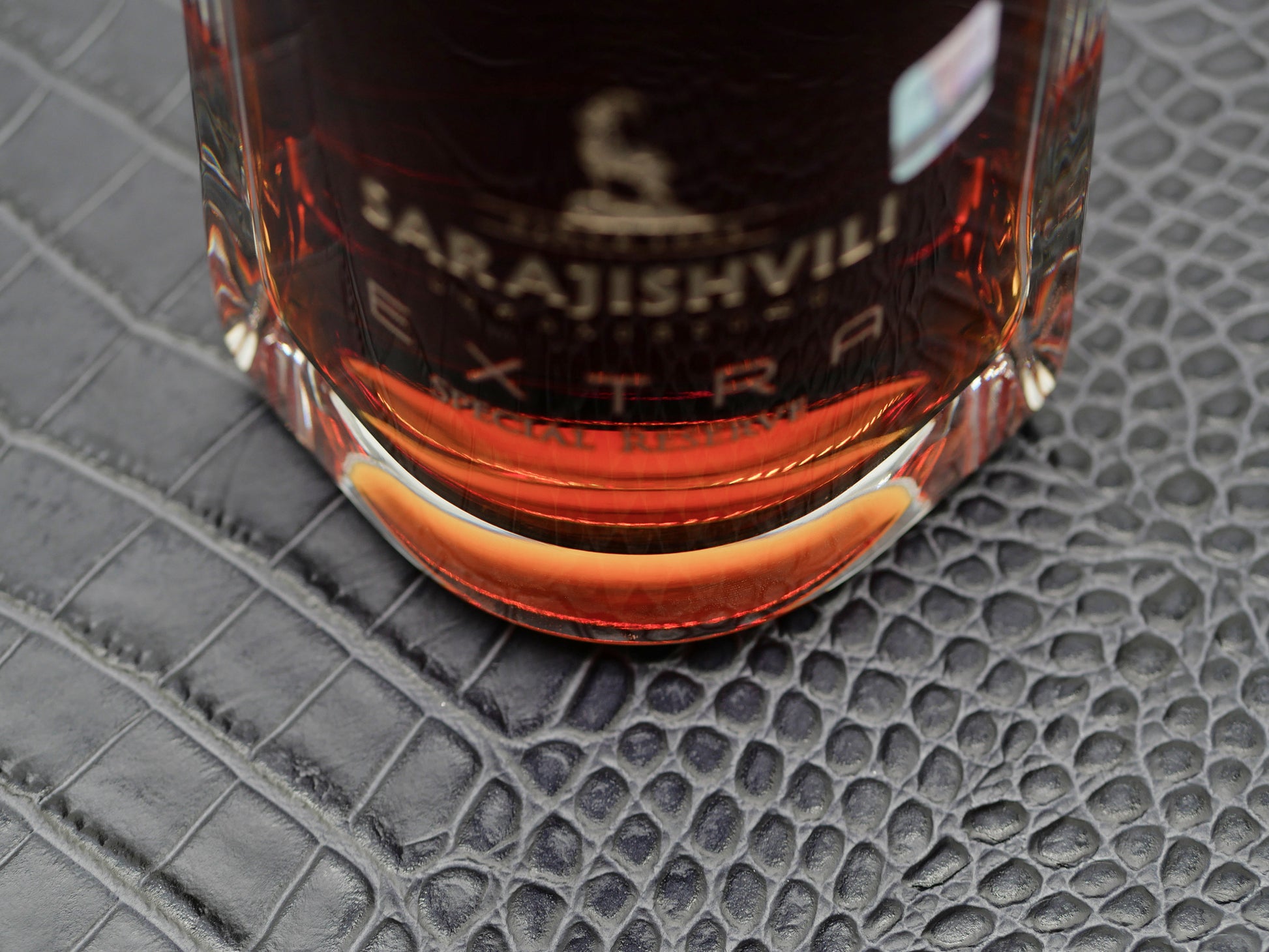 Close up of a glass bottle with amber liquid on a dark crocodile textured surface.