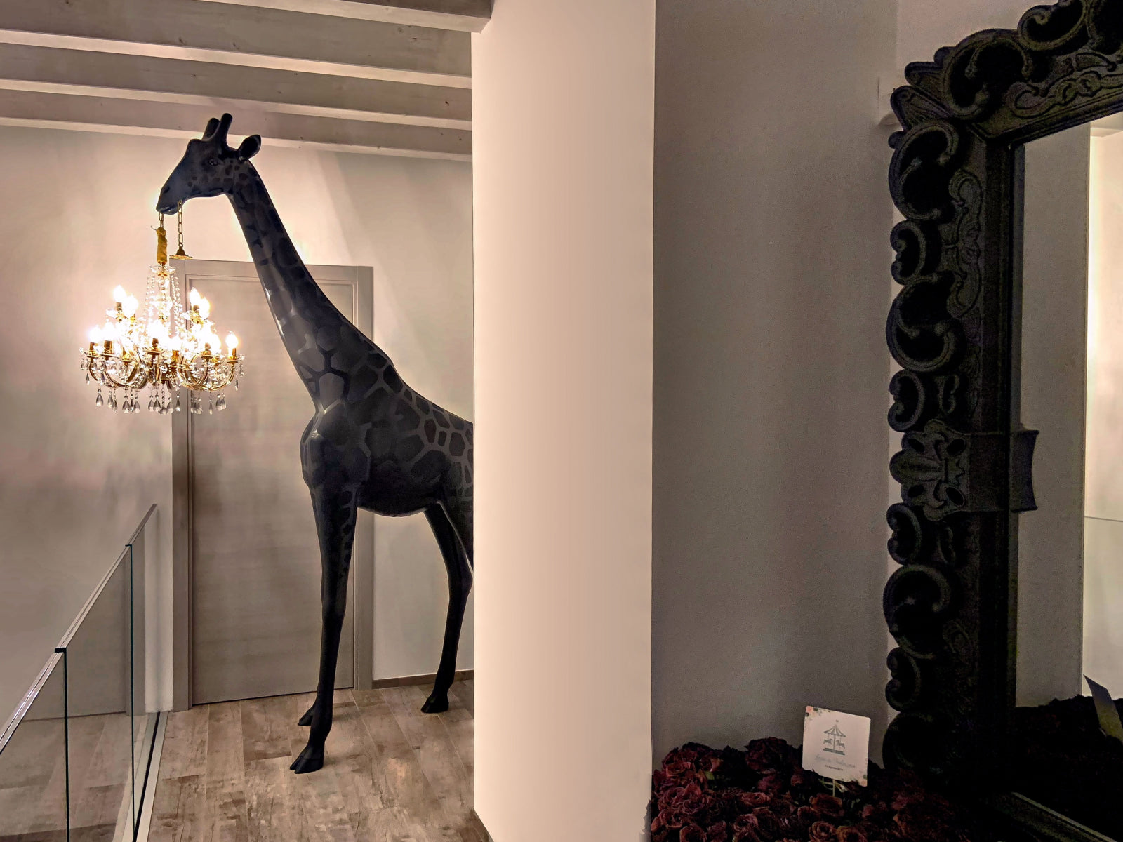 Giraffe in Love XS black sculpture stands in a modern interior with a gold Marie-Thérèse style chandelier hung around its neck.