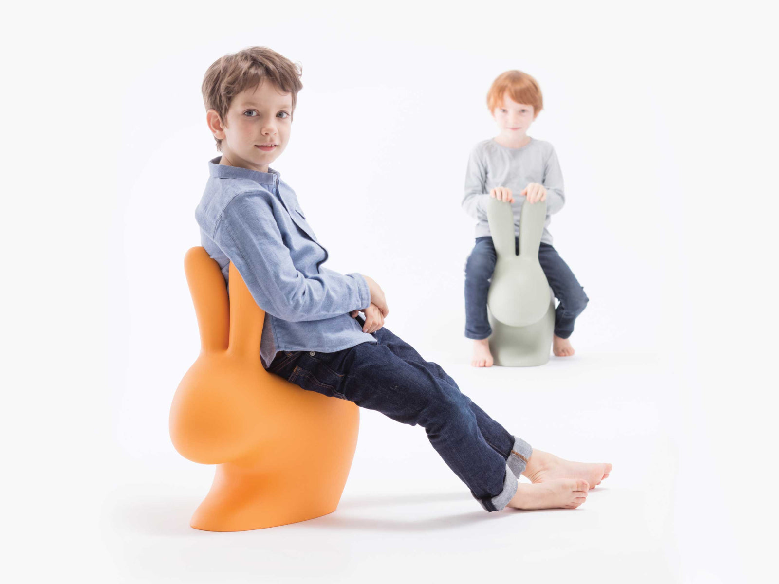 Two children sit on oversized Baby Rabbit Chairs in orange and pale green against a white background.