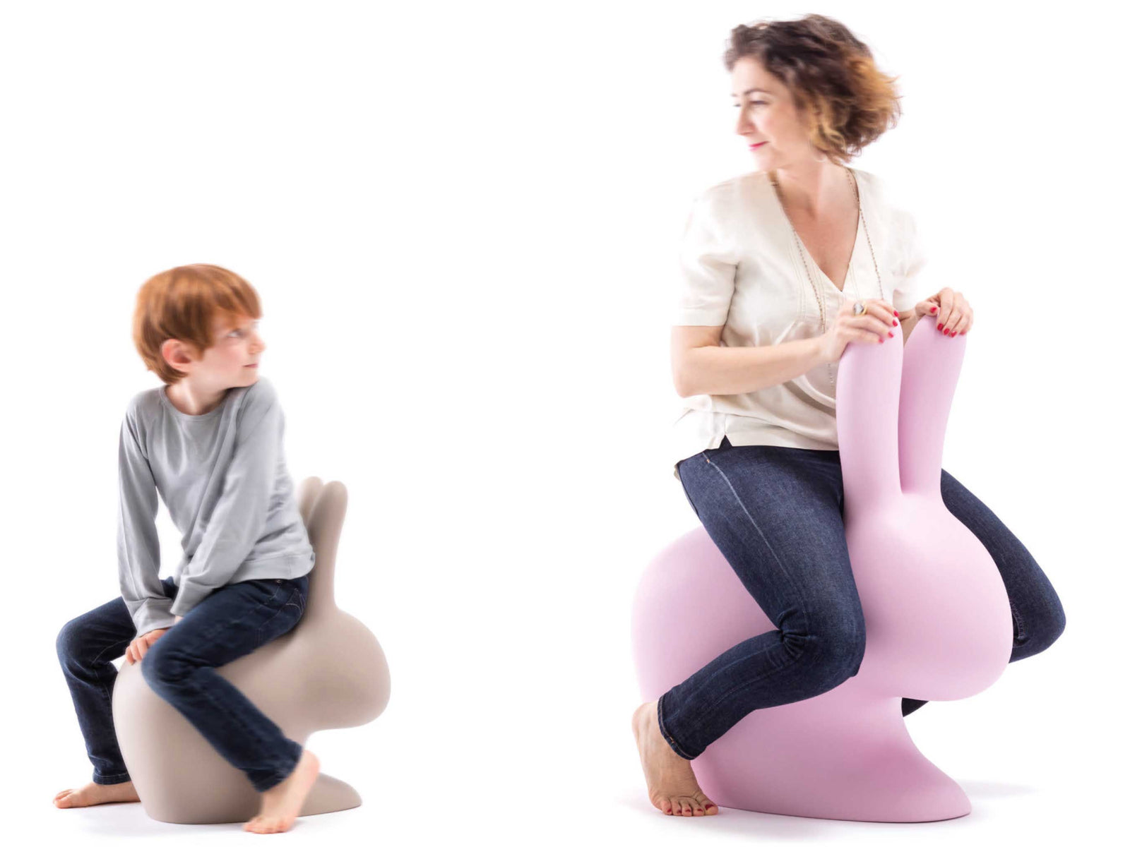 Child sits on beige Baby Rabbit Chair while a pink Baby Rabbit Chair supports a smiling woman in a bright studio