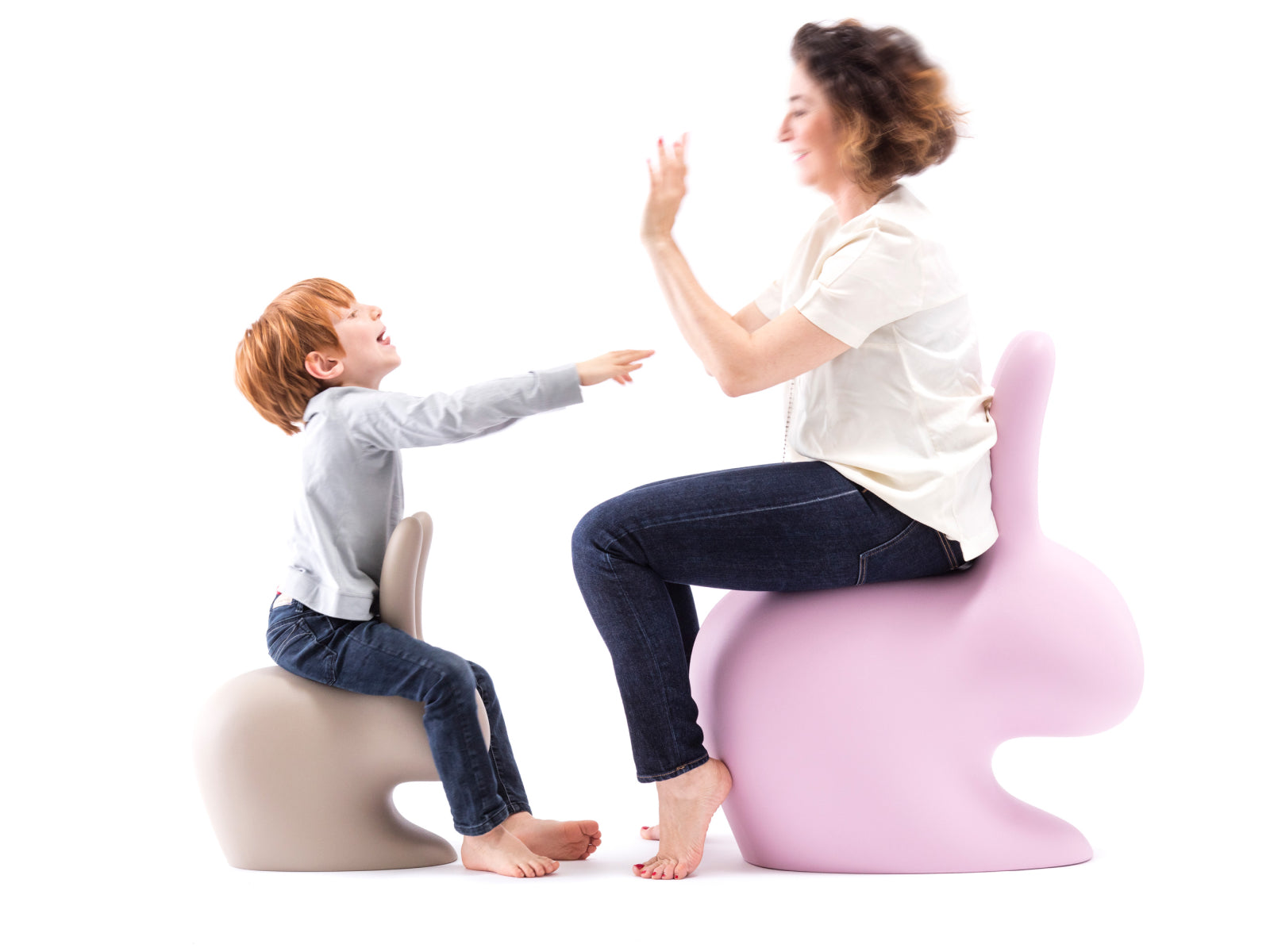 Child on beige Baby Rabbit Chair reaches toward a smiling woman on a pink Baby Rabbit Chair in a bright studio